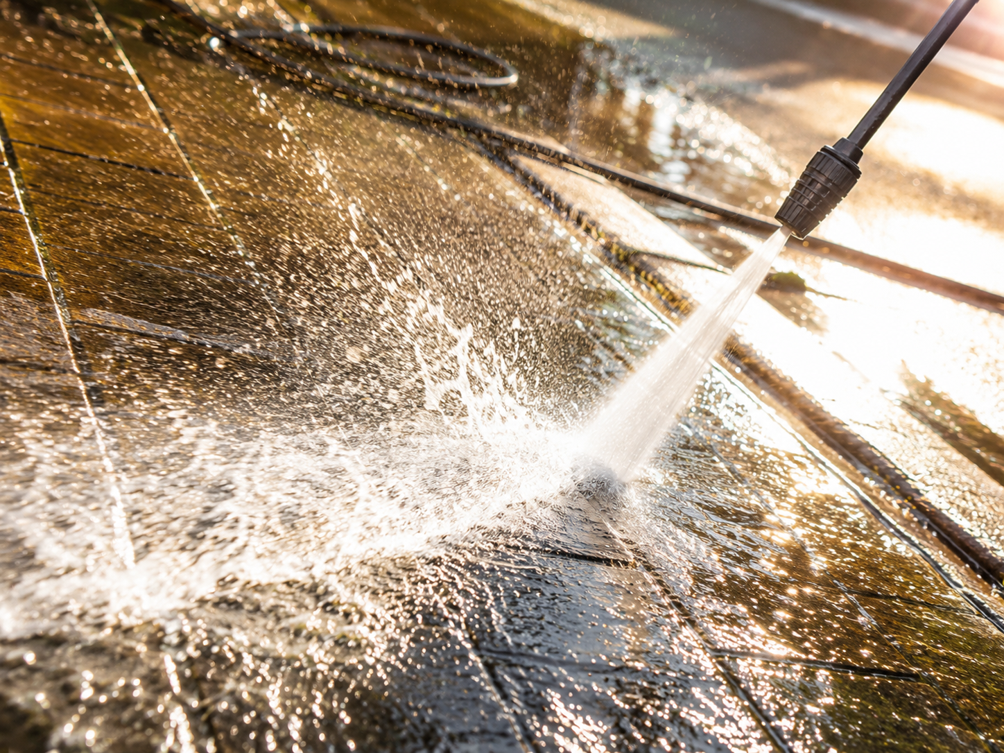 Close-up of a pressure washer spraying water on a wet outdoor surface, likely a driveway or pathway, with sunlight reflecting off the water droplets.