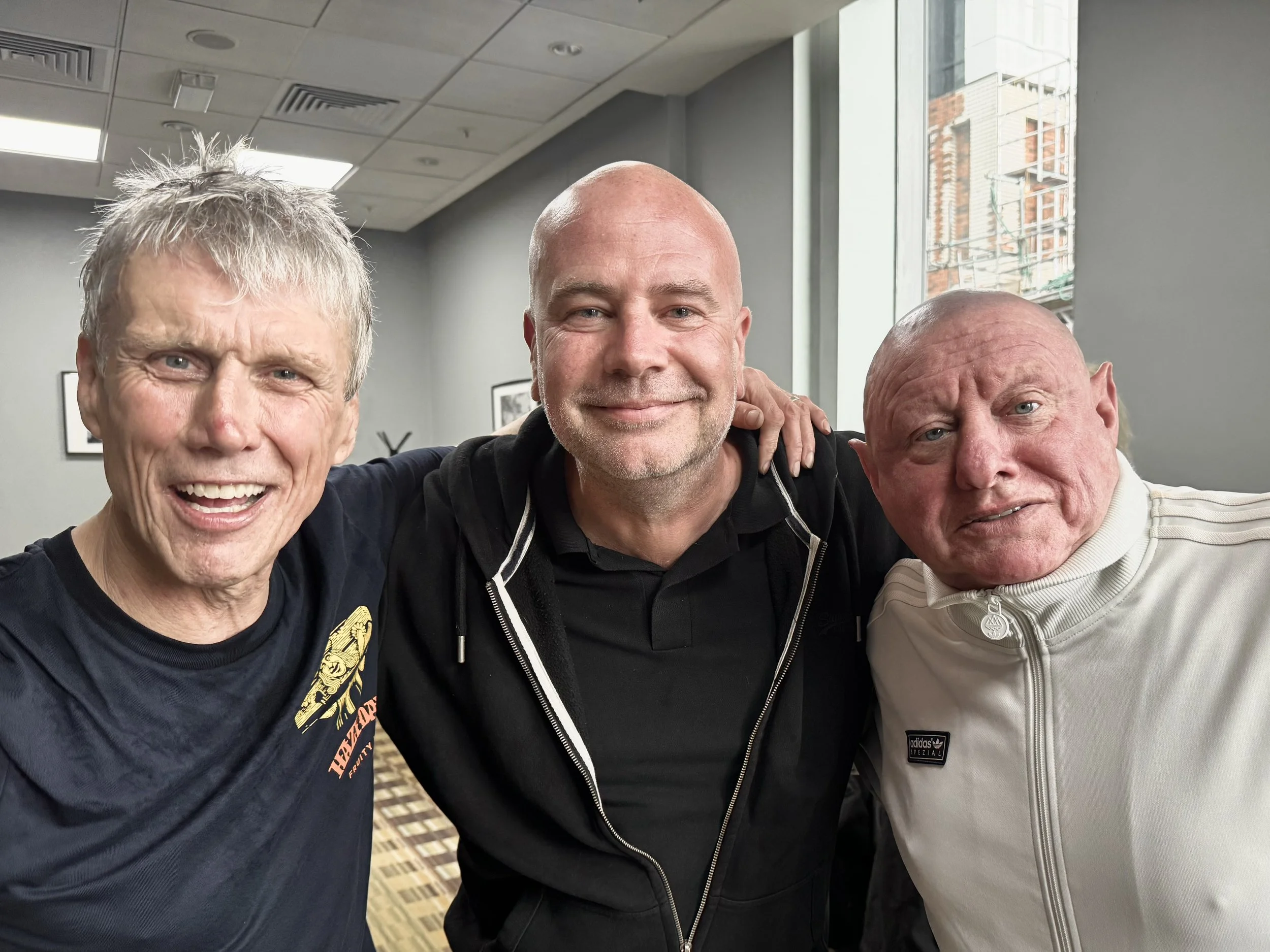 Three men smiling and posing closely together for a selfie in an indoor setting, with large windows showing a cityscape.