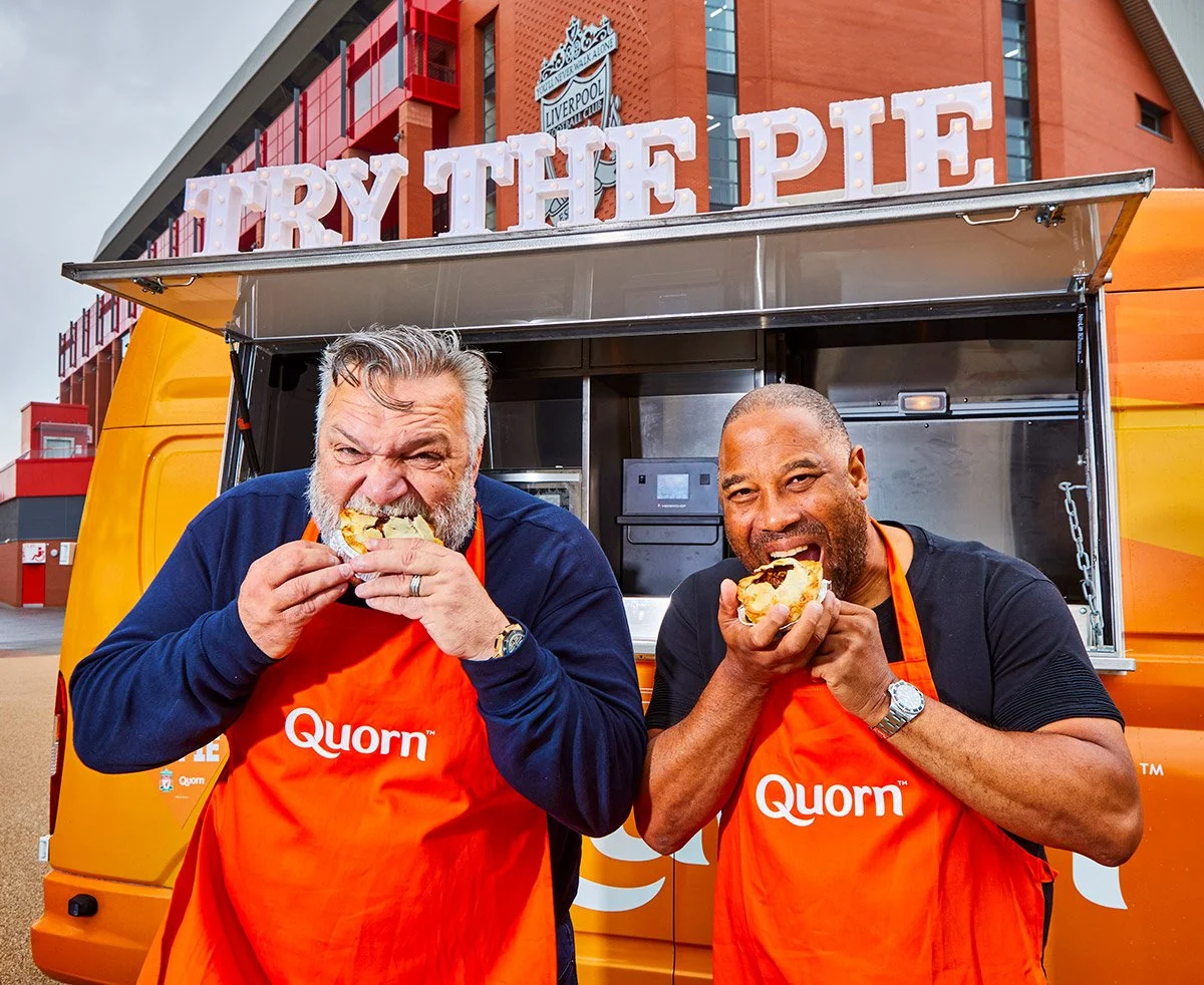 A PR photograph of two men wearing orange aprons with 'Quorn' logo enjoying eating burgers, standing in front of a food truck with 'TRY THE PIES' sign, and Liverpool Football Club logos on the building behind them.