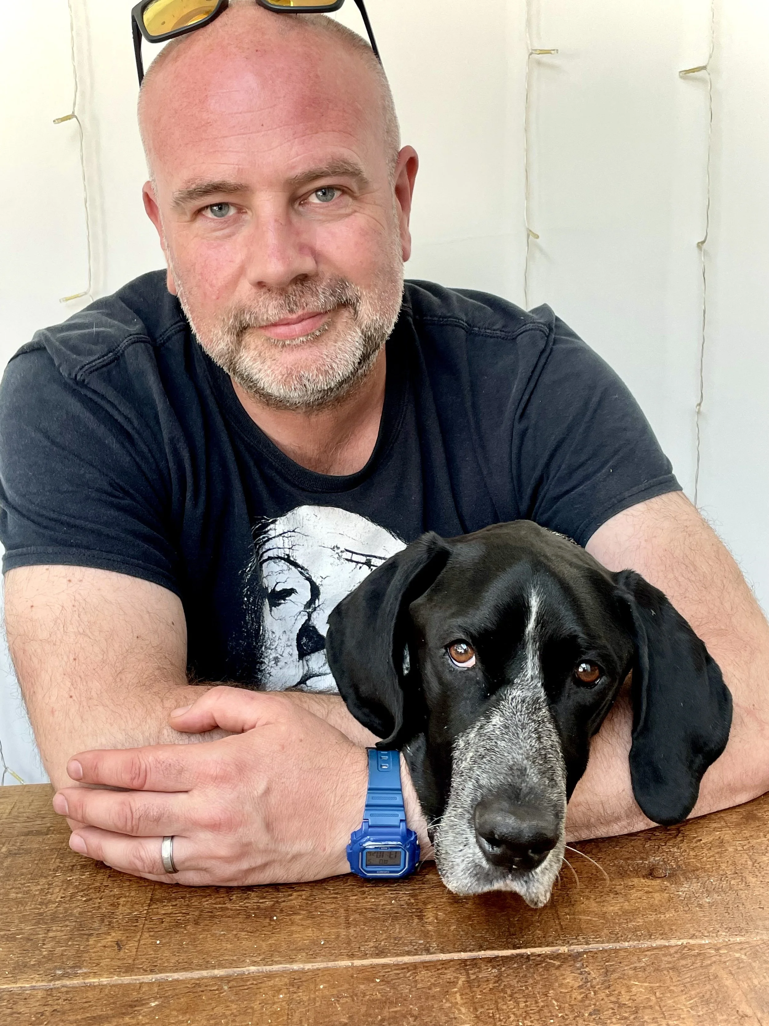 A man with a beard and light blue eyes, wearing a black t-shirt and a blue digital watch, sitting at a wooden table with a black and white dog resting its head on the table. The man is holding the dog with one arm, and his other arm is crossed on the table. The background has a white wall with string lights.