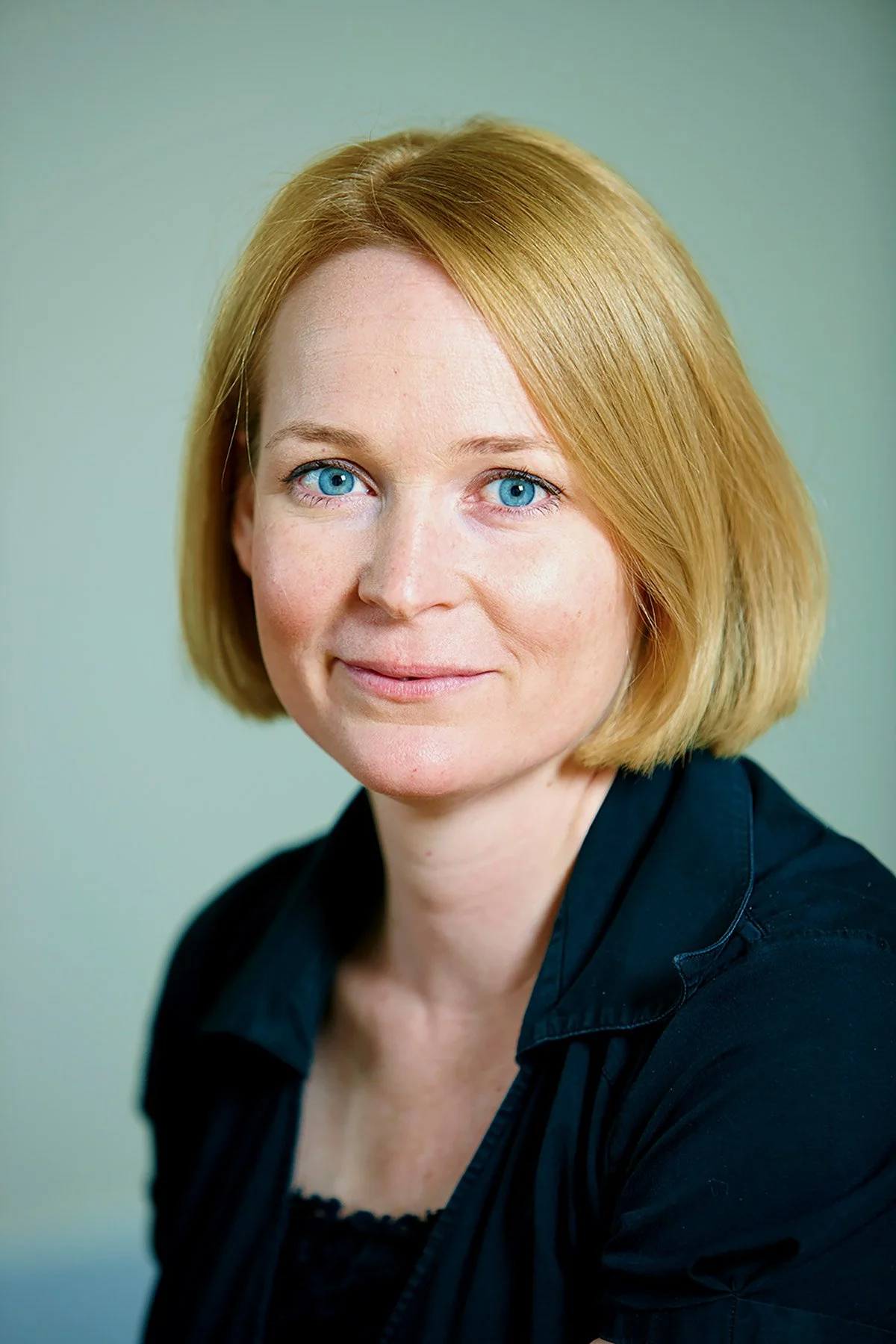 A portrait of a woman with short, blonde hair, blue eyes, and wearing a black top, against a light green background.