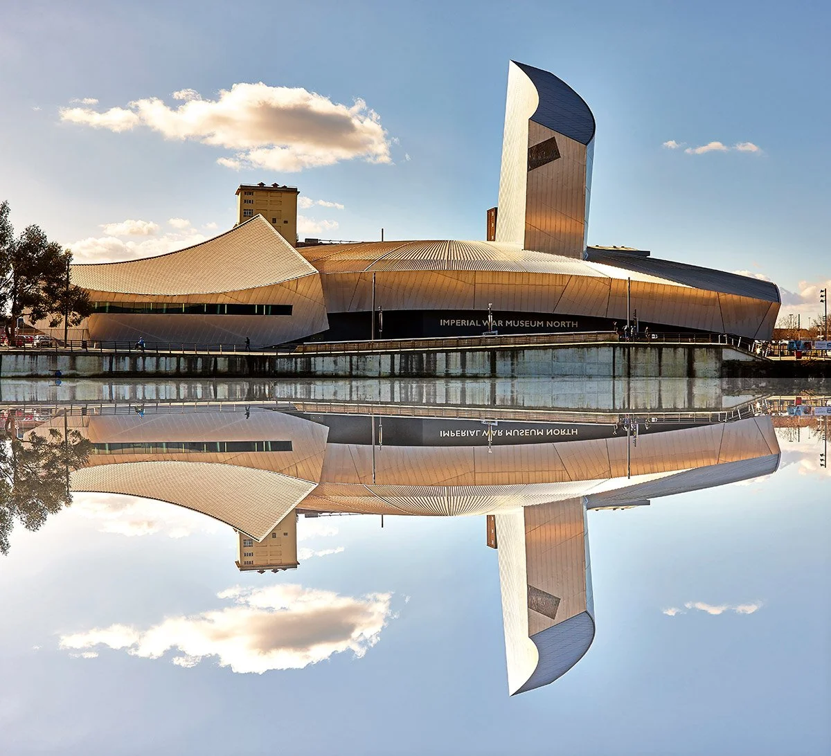 The Imperial War Museum North building in Manchester, UK, reflecting on a body of water during the day with a partly cloudy sky.