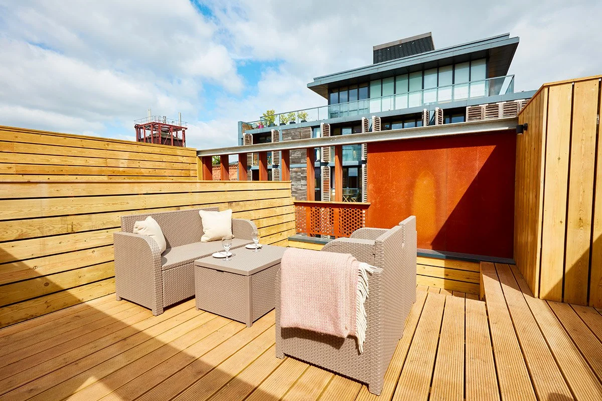 Outdoor balcony with wicker furniture, including a sofa and two armchairs, a small table with glasses, and a pink throw blanket, surrounded by wooden fencing and overlooking modern buildings with a partly cloudy sky.