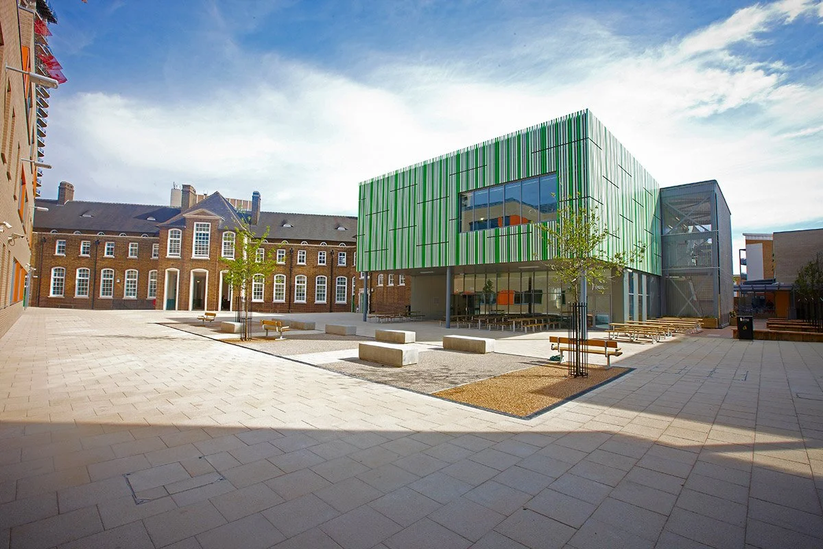 A modern urban plaza with paved walkways, small trees, benches, and a mix of old brick and contemporary glass buildings under a partly cloudy sky.