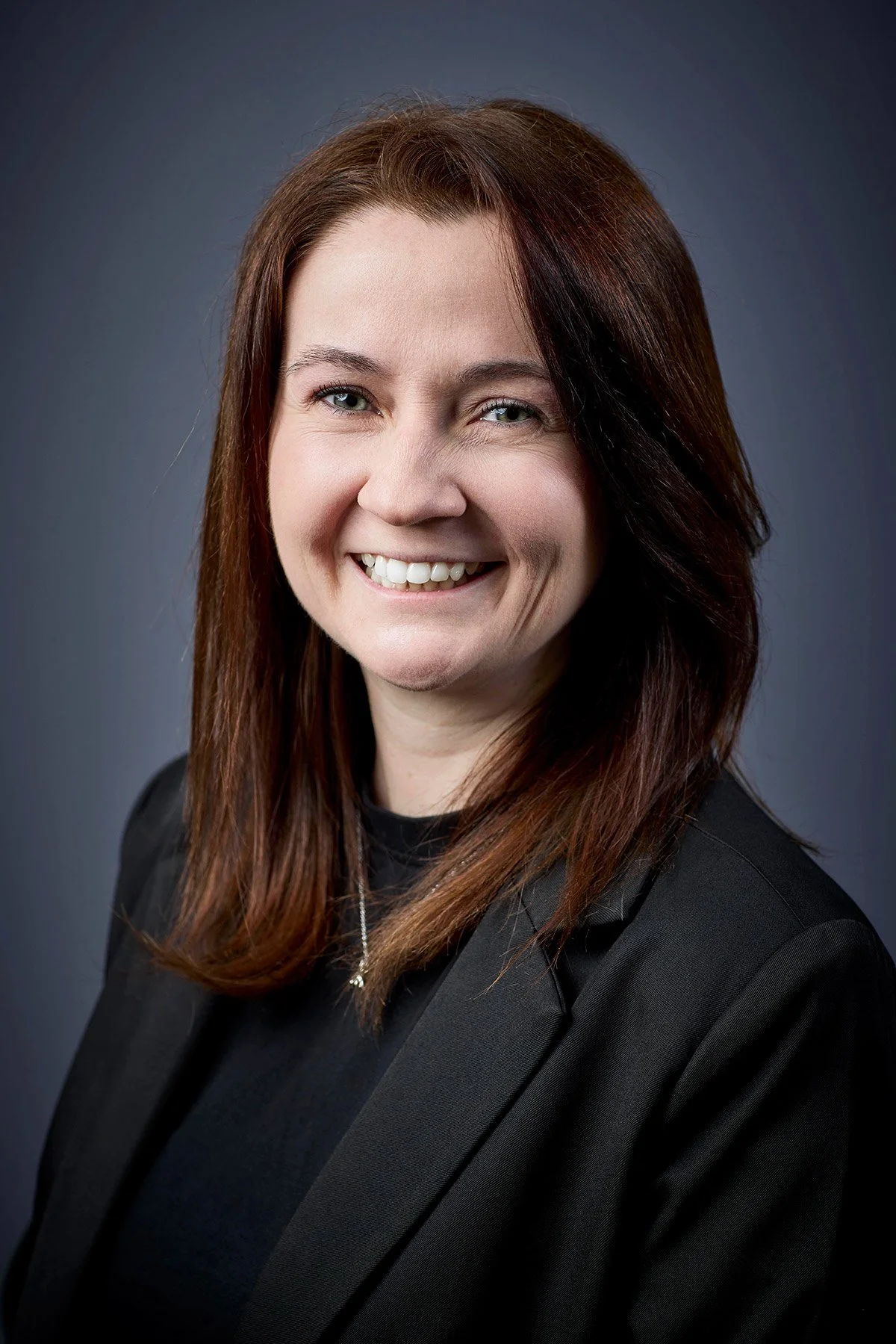 Professional headshot of a woman with shoulder-length reddish-brown hair, smiling, wearing a black blazer and a necklace, against a dark gray background.