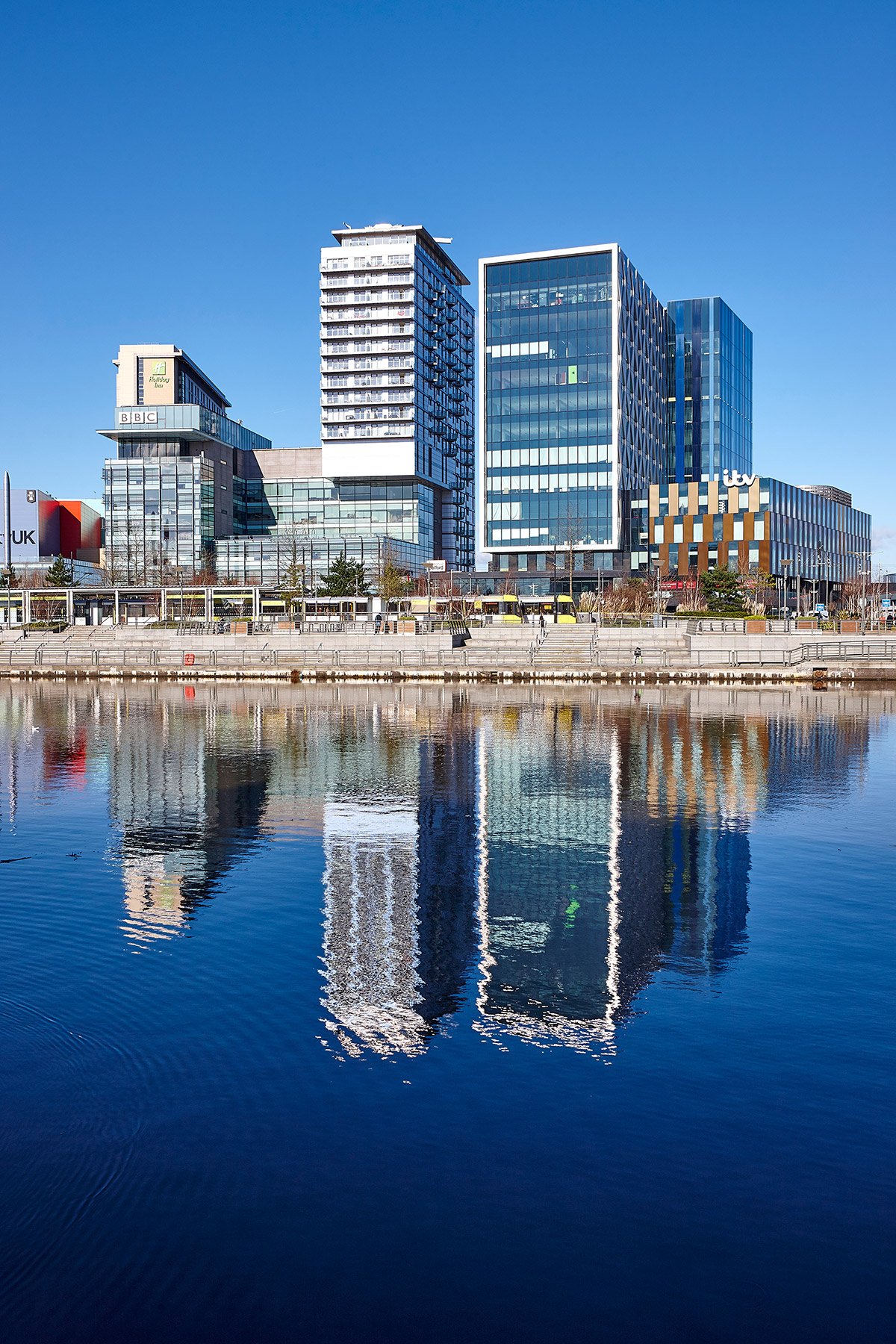 Modern cityscape with tall glass buildings reflected in a river, under a clear blue sky.