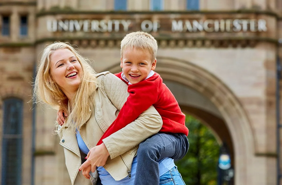 A woman giving a piggyback ride to a boy in front of the University of Manchester gate, both smiling and laughing.