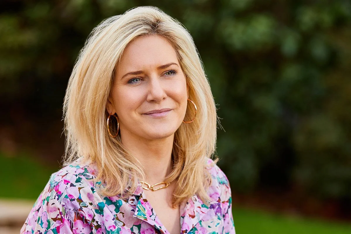 A woman with blonde hair, blue eyes, and a gentle smile, wearing a colorful floral blouse, gold hoop earrings, and a gold chain necklace outdoors with a green blurred background.