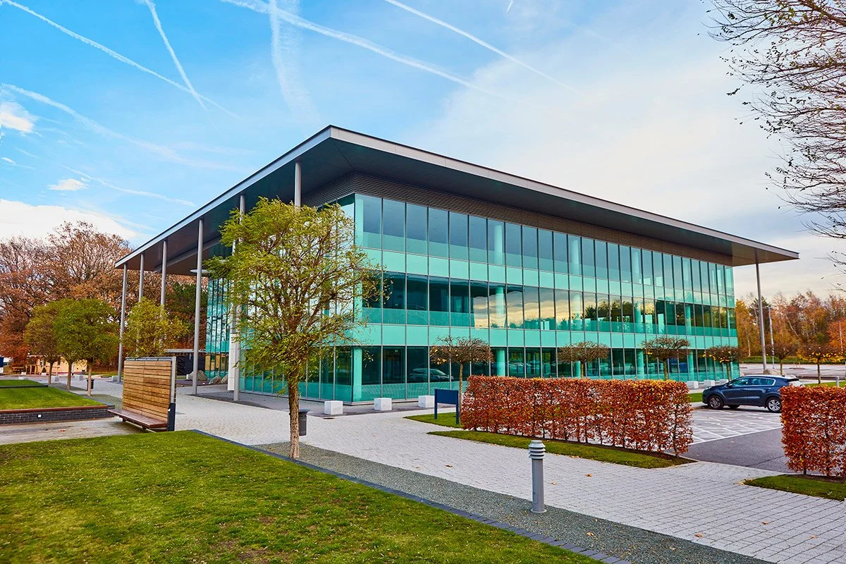 A modern glass office building with a sloped roof, surrounded by trees, bushes, and a parking lot under a partly cloudy sky.