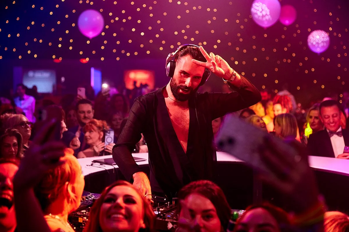 A DJ with headphones at a lively party, making a peace sign with his hand, surrounded by a crowded dance floor with colorful lights and dancing people.
