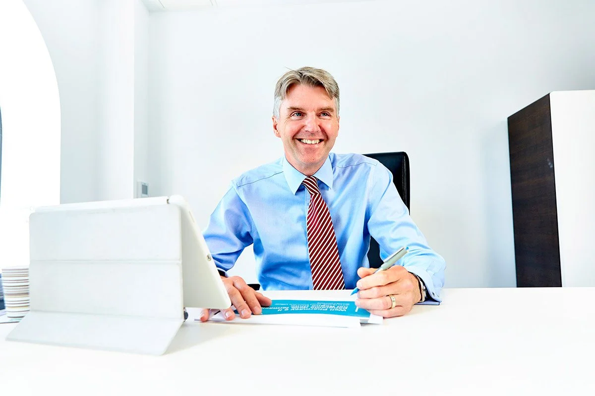 A smiling man in a blue dress shirt with a red and white striped tie sitting at a desk in an office, holding a pen, with a tablet and documents in front of him.