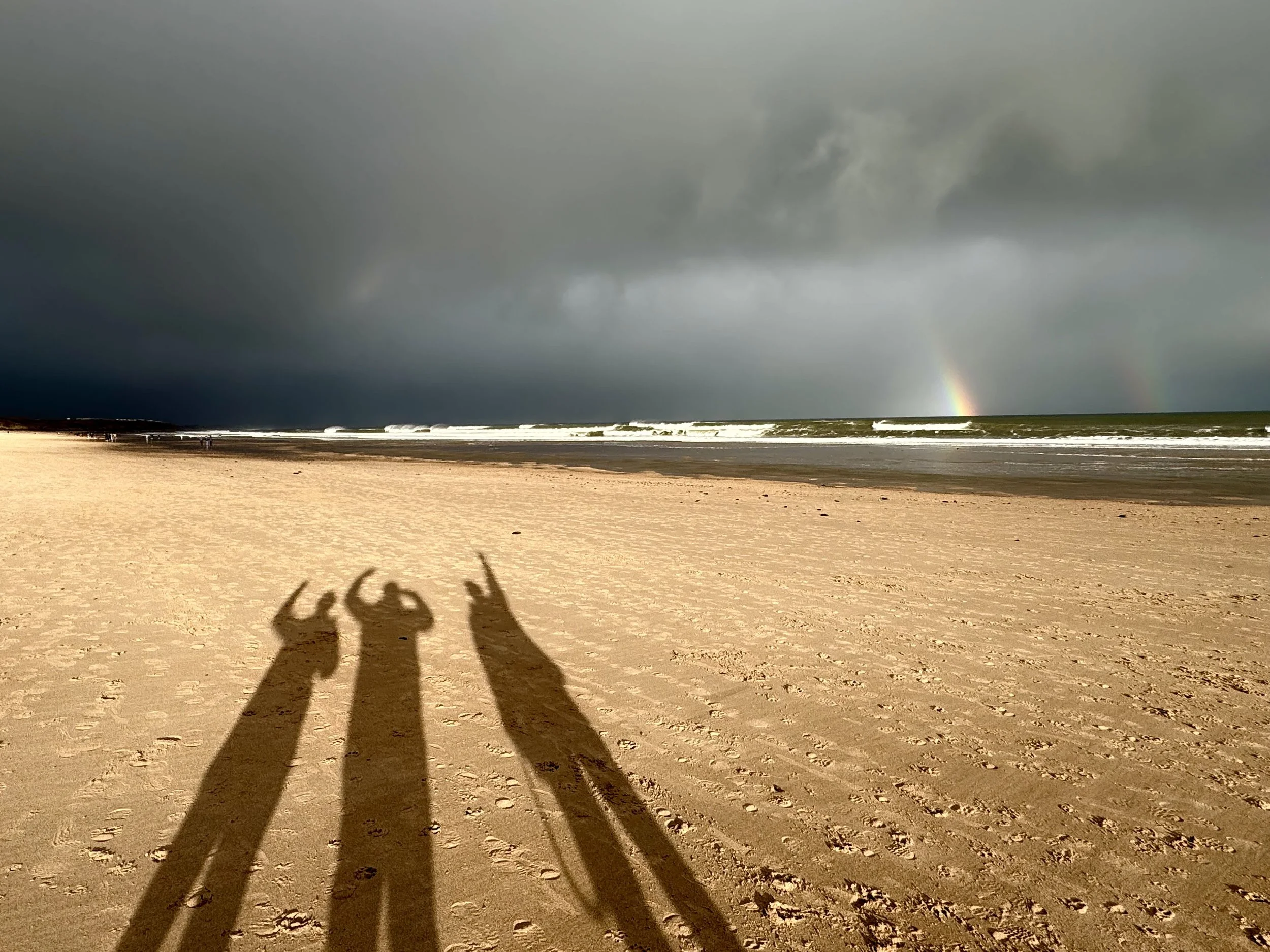 Shadow of two people taking a photo on a sandy beach with dark storm clouds and a rainbow in the sky.