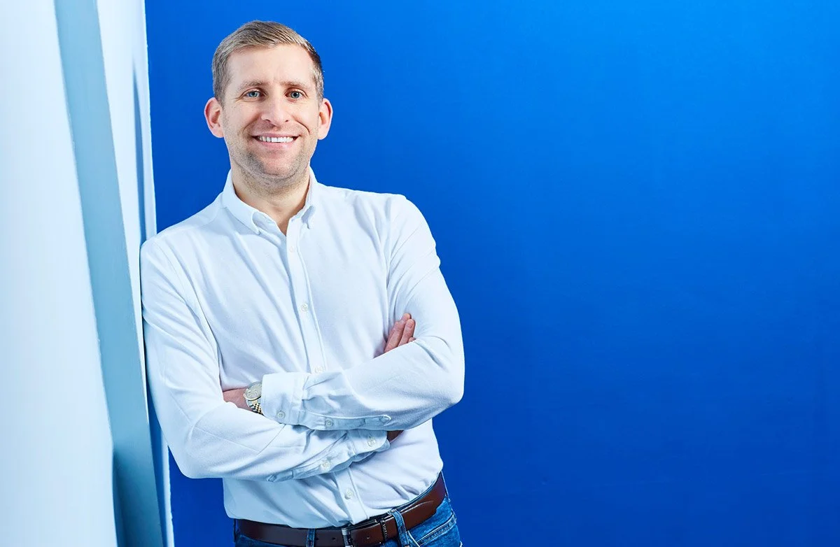 A corporate portrait of a smiling man in a white dress shirt with arms crossed, standing against a blue wall.