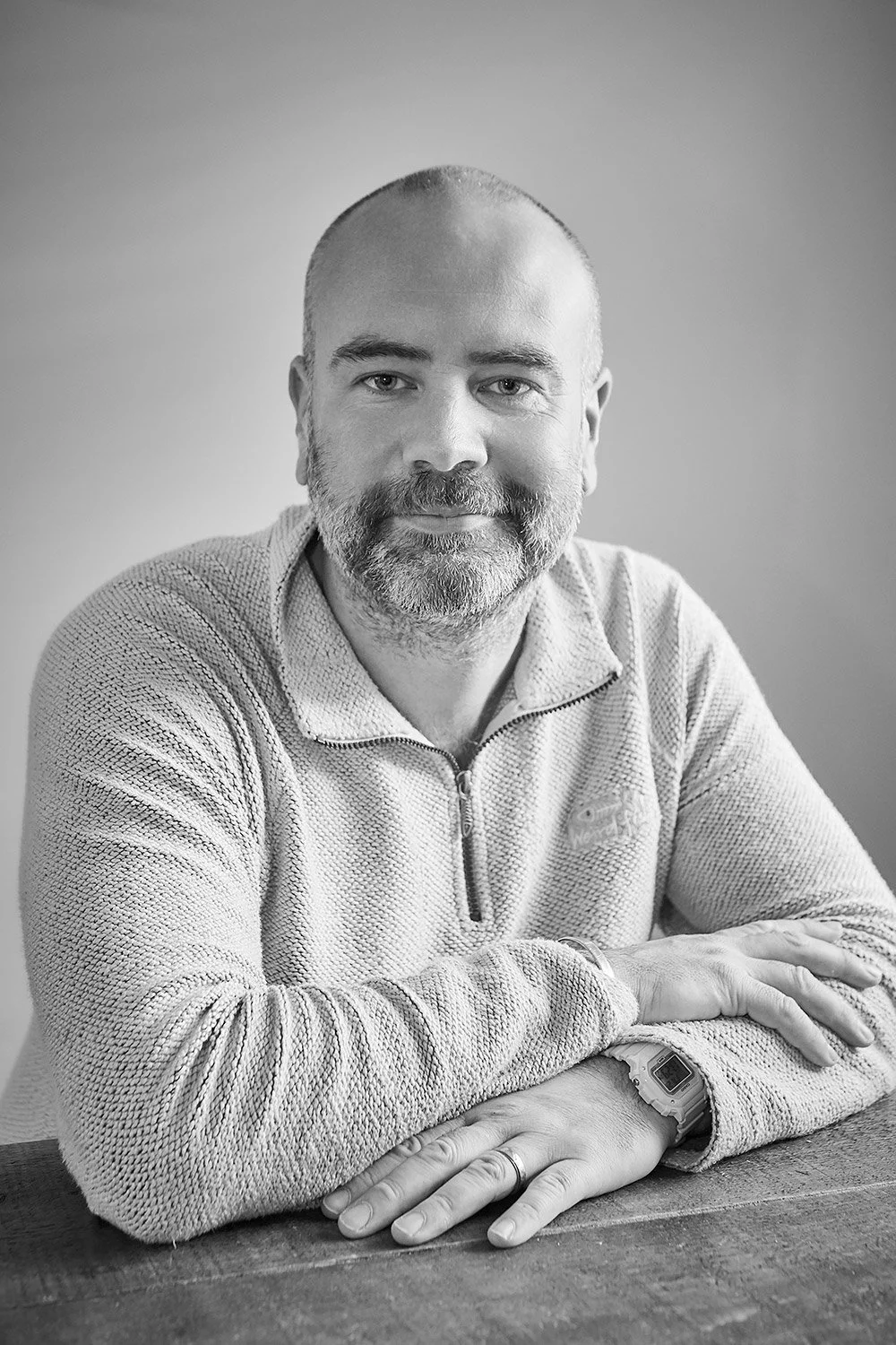 Black and white photo of a man with a beard and short hair, sitting at a table with his arms crossed, wearing a textured quarter-zip sweater and a digital watch.