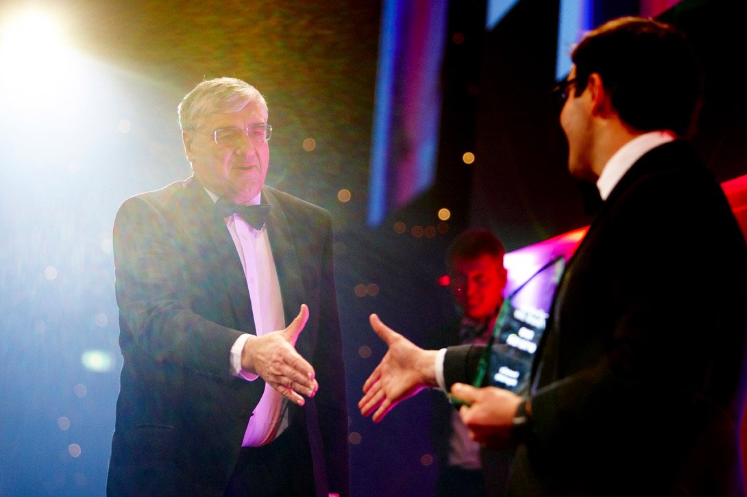 Two men in tuxedos exchanging handshakes at a formal event with colorful lighting and blurred background.