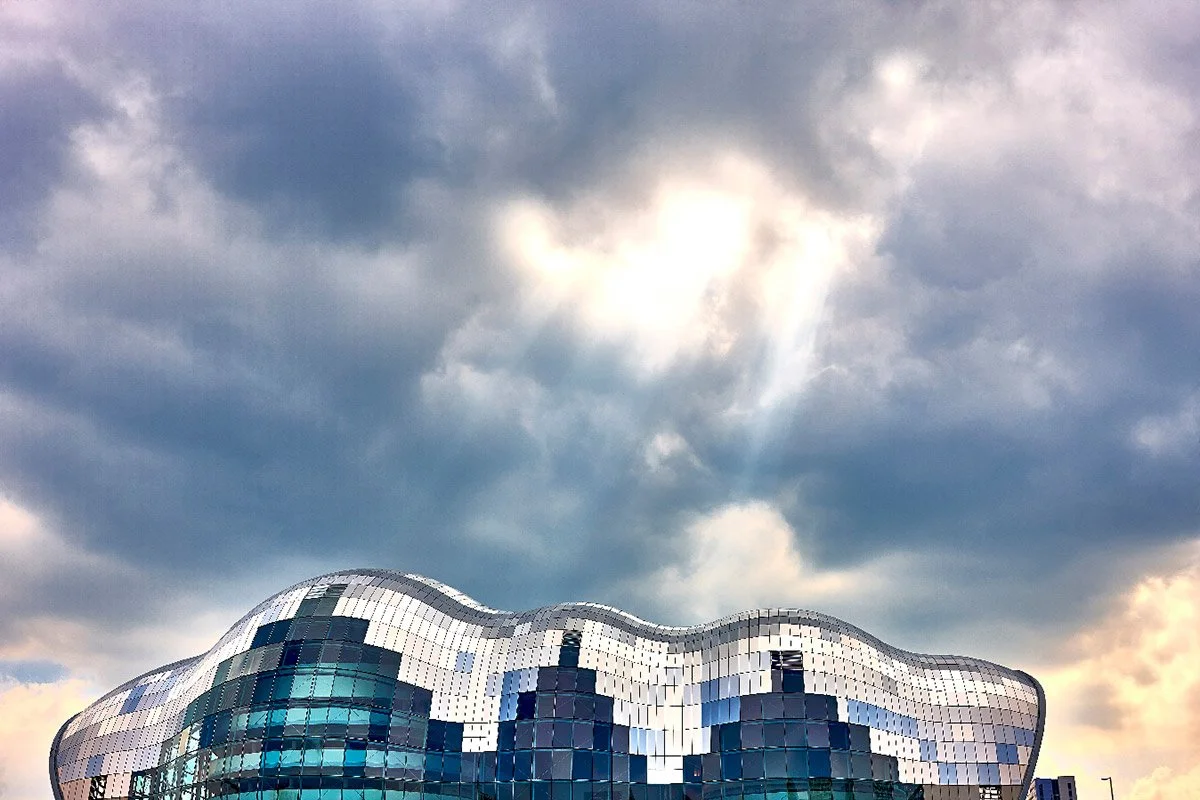 A modern photography of a building with a wavy, reflective glass facade against a cloudy sky.