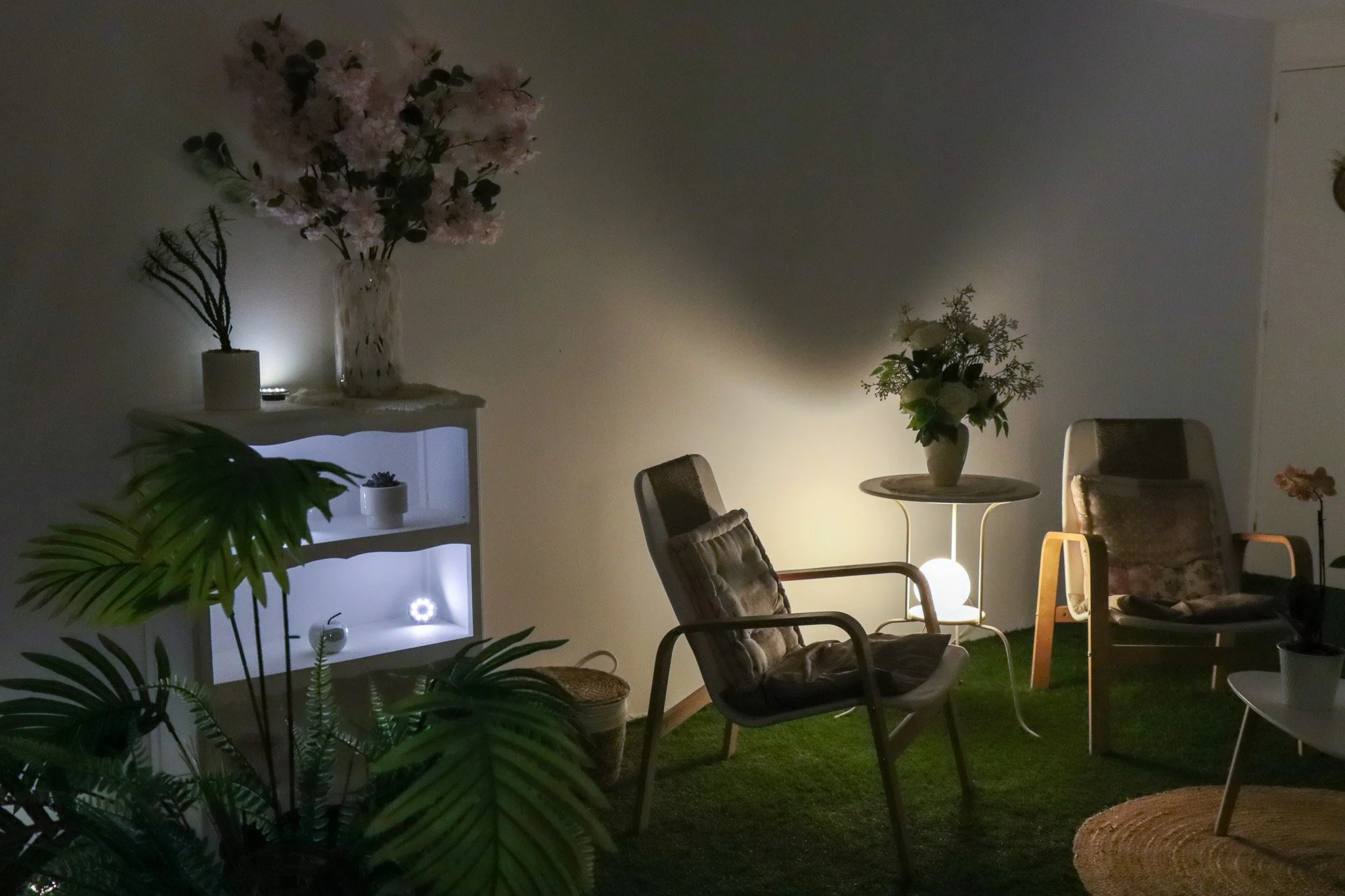 Un coin salon intérieur avec des chaises et des tables, plantes vertes, étagère avec des décorations, et des lampes d'ambiance. La pièce est éclairée par des lumières douces.