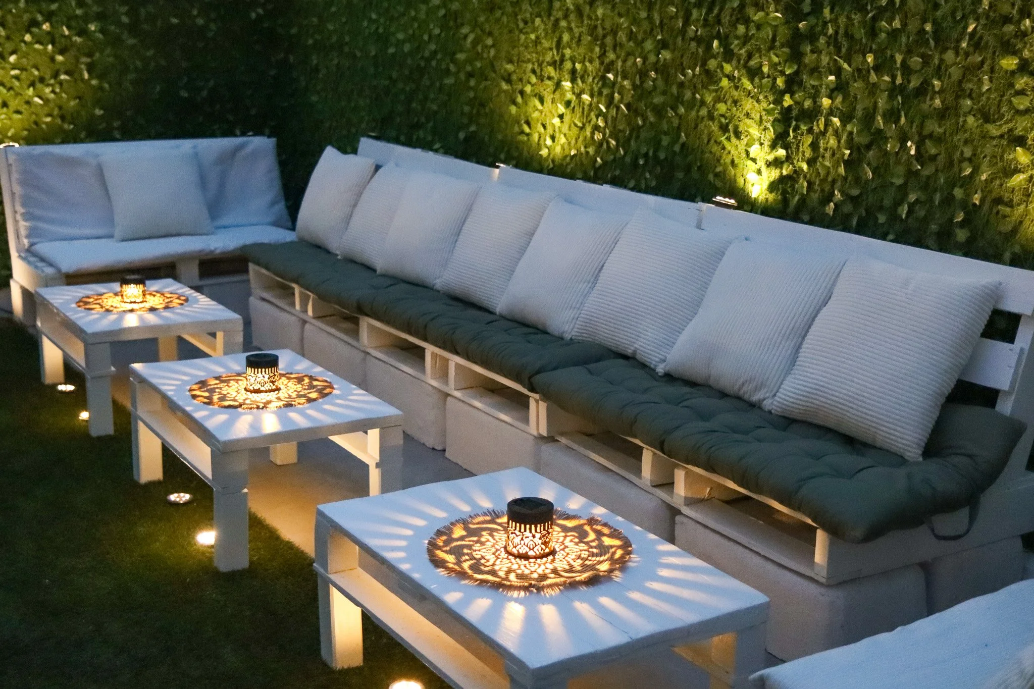 Une banquette en bois blanc avec des coussins gris et blancs est placée devant un mur végétal éclairé à la nuit. De petites tables blanches avec des lampes décoratives en métal montrent des motifs d'ombres sur leur surface.