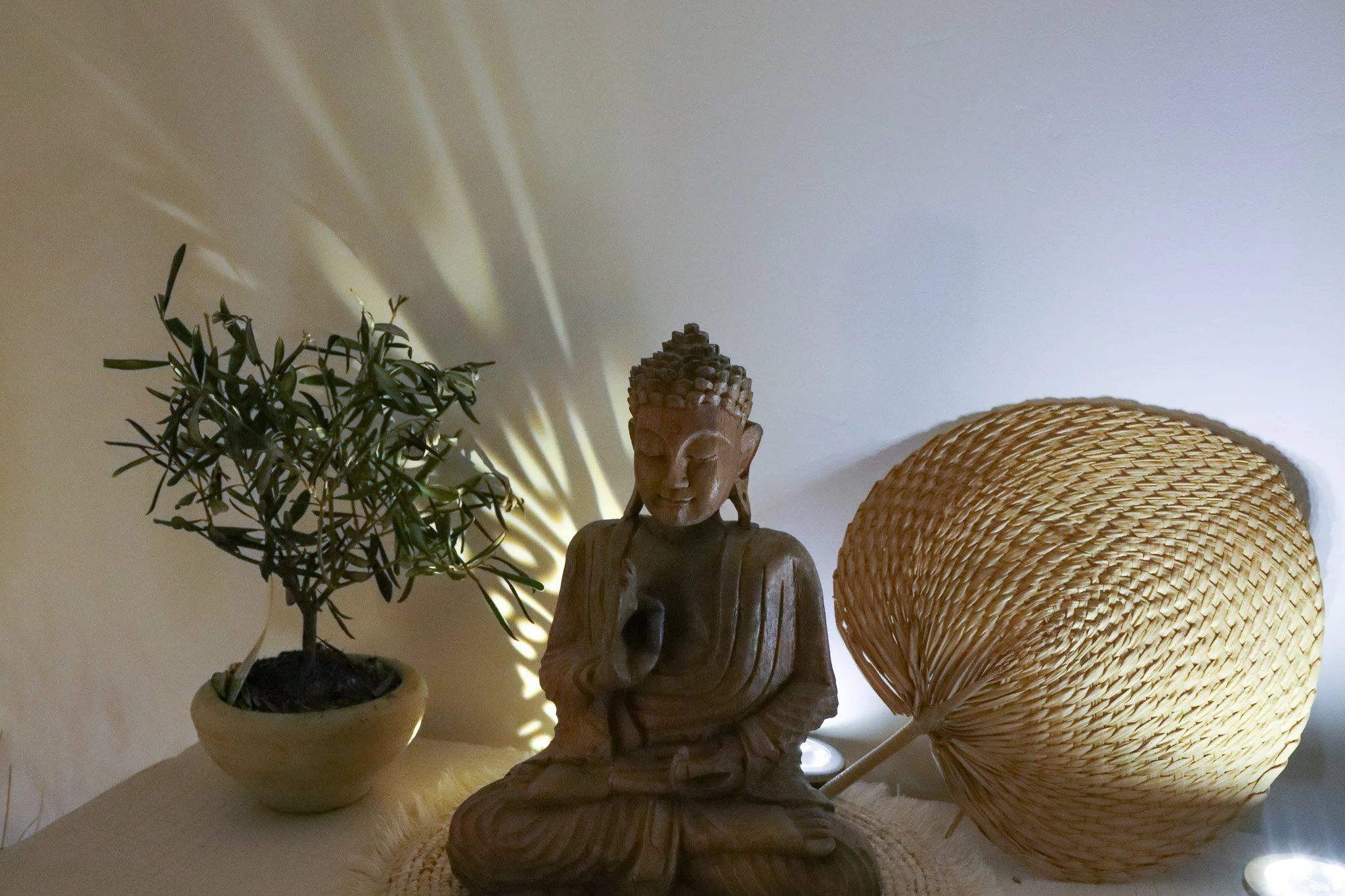 Statue en bois d'une figure bouddhiste assise, entourée d'une plante en pot à gauche et d'un chapeau en osier à droite, avec un éclairage qui crée un jeu d'ombres sur le plafond.