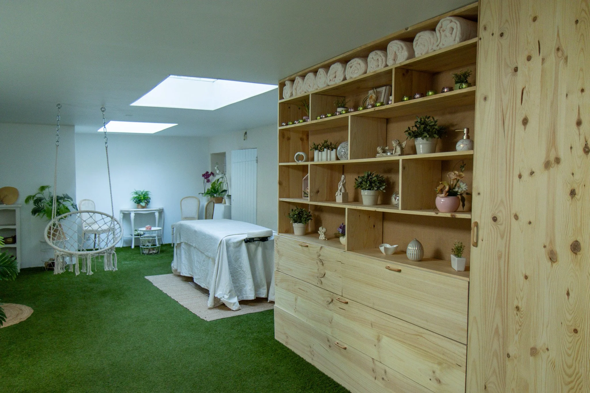 Intérieur d'une pièce avec un grand meuble en bois rempli de plantes, décorations et serviettes blanches soigneusement roulées. La pièce est lumineuse avec deux puits de lumière au plafond, un coin salon avec une table et un canapé à motifs blancs et des fauteuils, le sol est recouvert de pelouse artificielle.