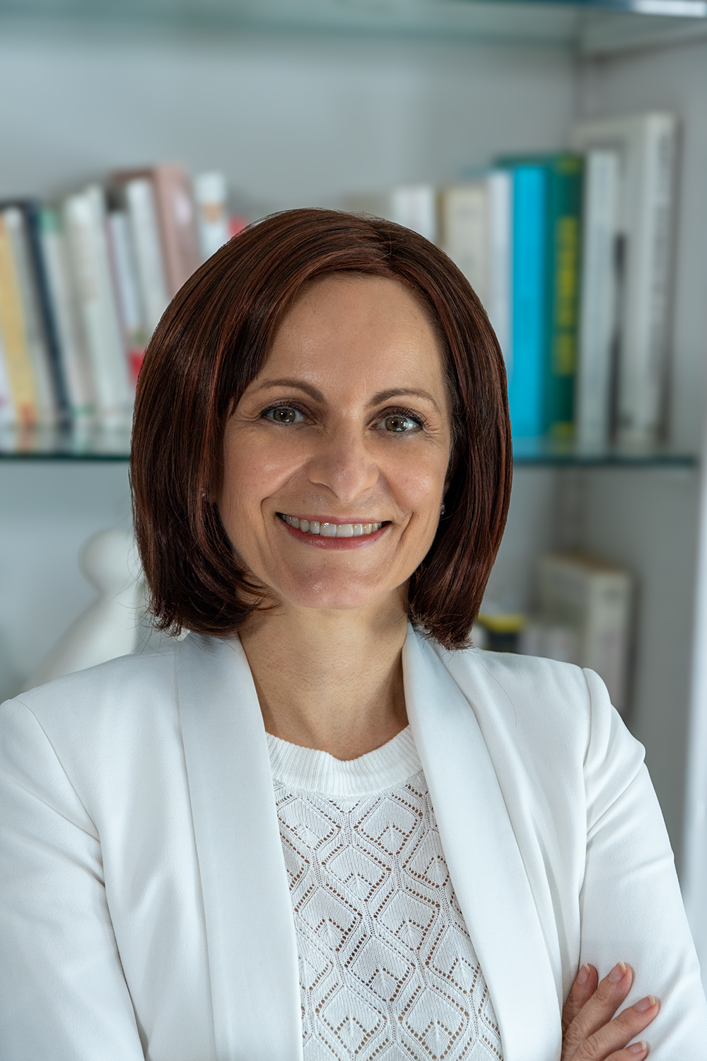 Femme souriante en veste blanche dans un bureau avec des étagères remplies de livres.