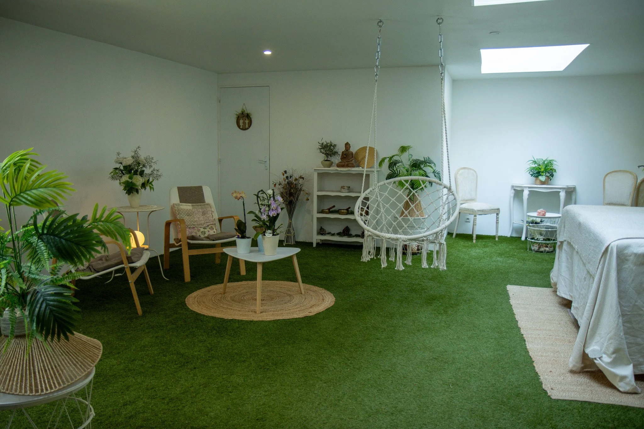 Intérieur lumineux avec des meubles en bois, des plantes vertes, un fauteuil, une table ronde avec une plante en pot, un fauteuil suspendu en macramé, des étagères avec des objets décoratifs, un canapé couvert d'une nappe, un tapis en osier, un tapis beige et un plafond avec un puits de lumière.
