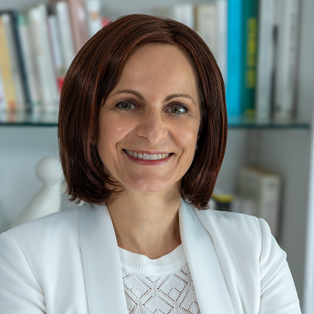 Une femme souriante avec les cheveux bruns, portant un blazer blanc, dans un bureau intérieur.