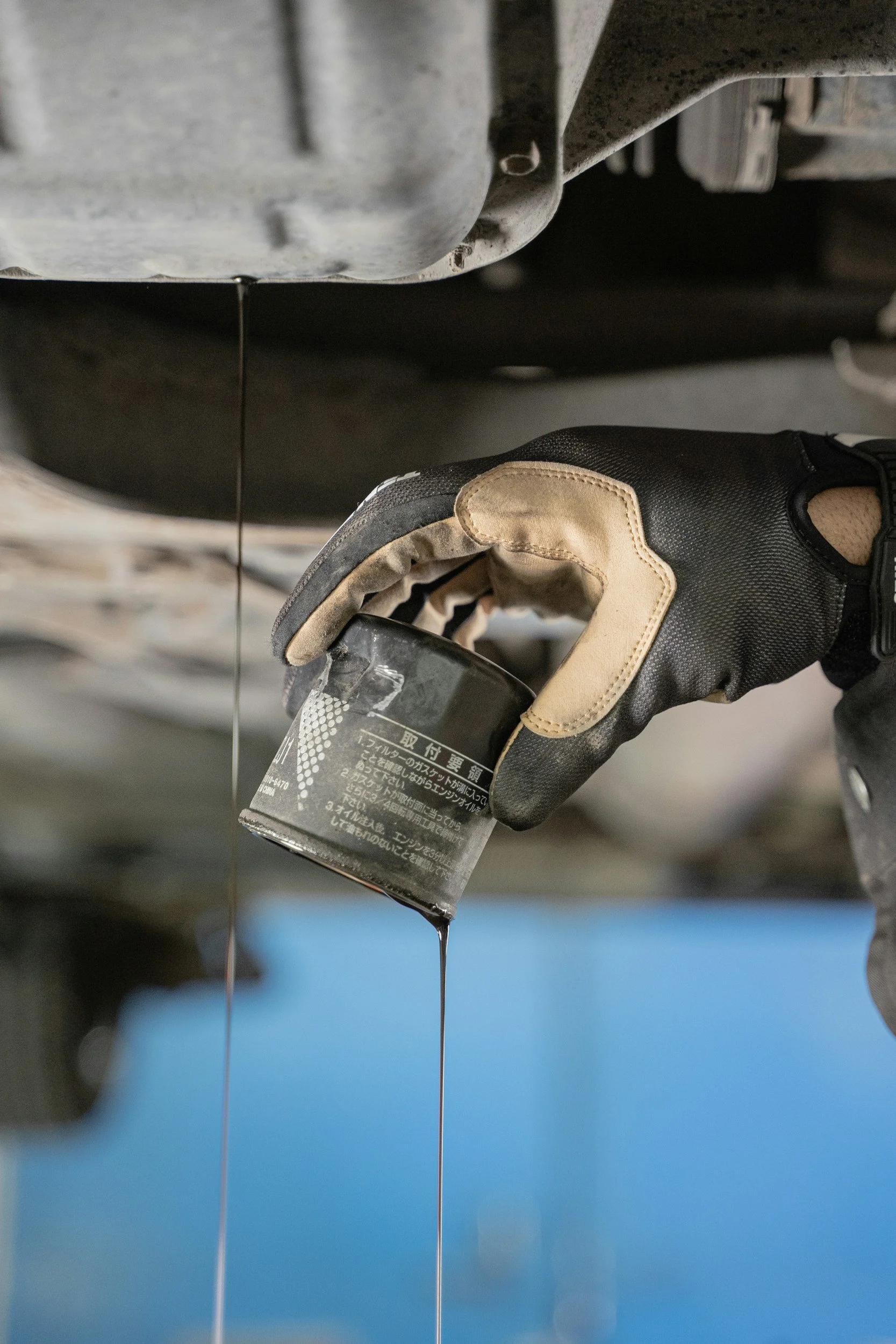 A person wearing black and beige work gloves holding a black bottle underneath a vehicle, with black engine oil dripping from the oil pan. The focus is on the oil stream and the person's gloved hand.