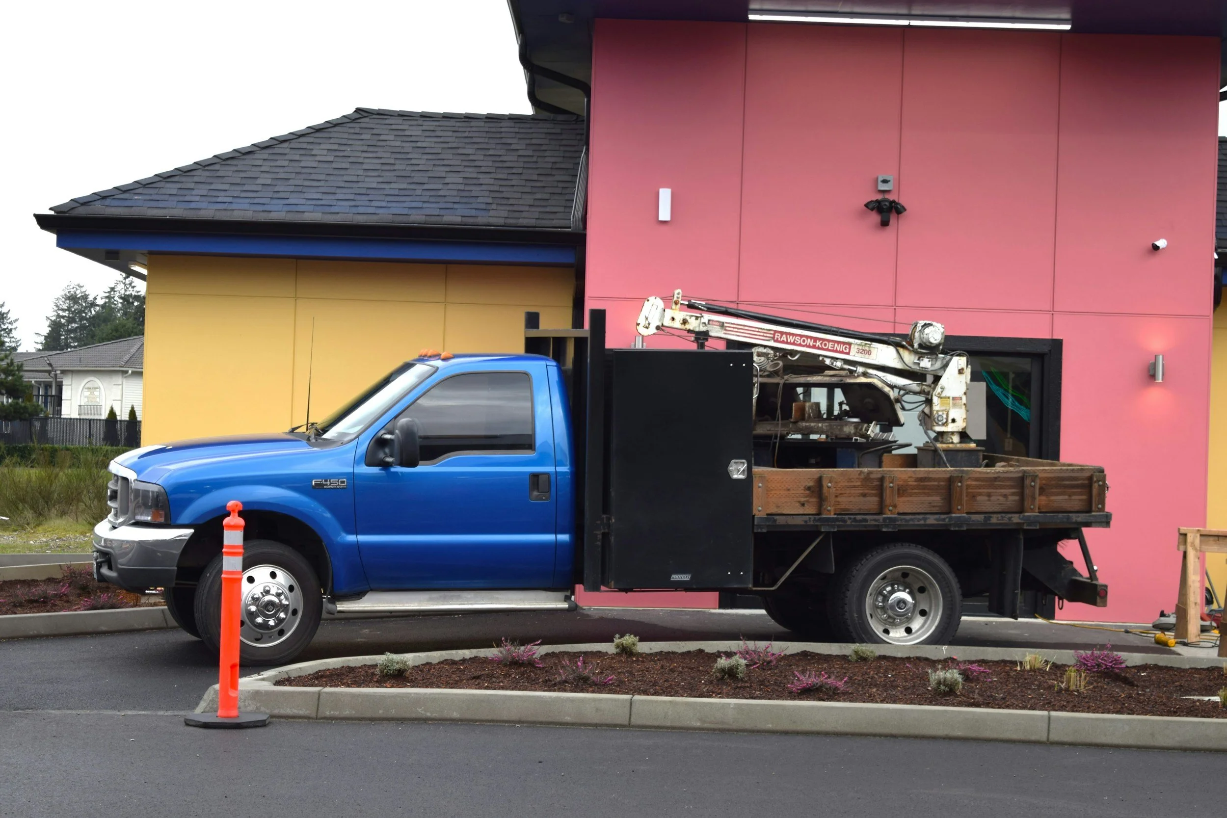 A blue truck with an attached flatbed holding construction equipment parked in front of a partially painted building with pink and yellow walls. Construction work is ongoing.