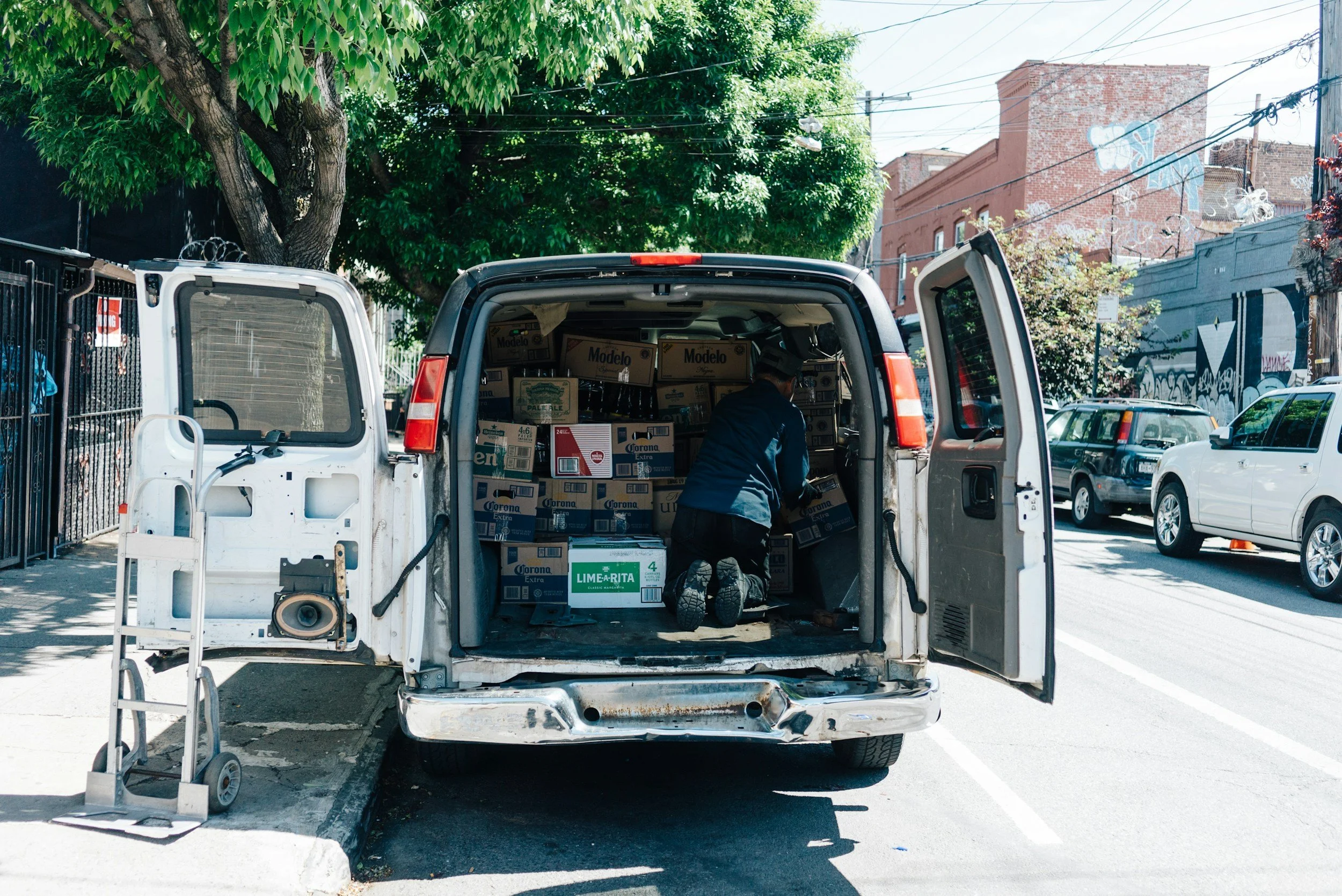 A white van with its back doors open, revealing boxes of beer inside. A person is kneeling inside the van, unpacking or organizing the boxes. The scene is outdoors on a city street with parked cars and buildings in the background, trees overhead, and power lines above.
