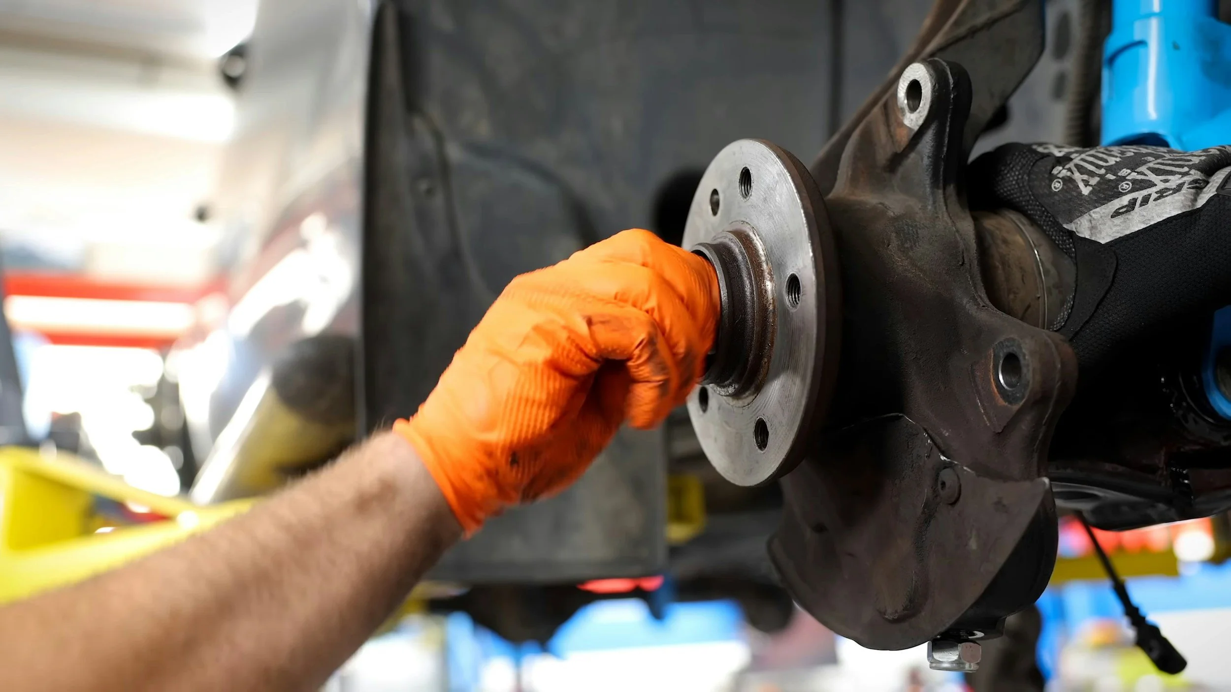 A mechanic wearing orange gloves working on a car's brake rotor in an automotive repair shop.