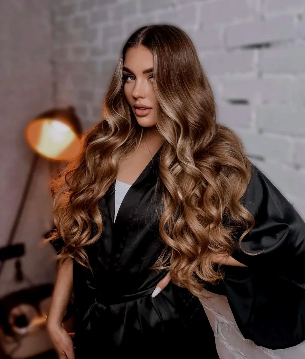 A young woman with long, wavy, light brown hair posing in front of a gray brick wall, wearing a black satin robe and white lace nightgown, with soft lighting and a blurred lamp in the background.