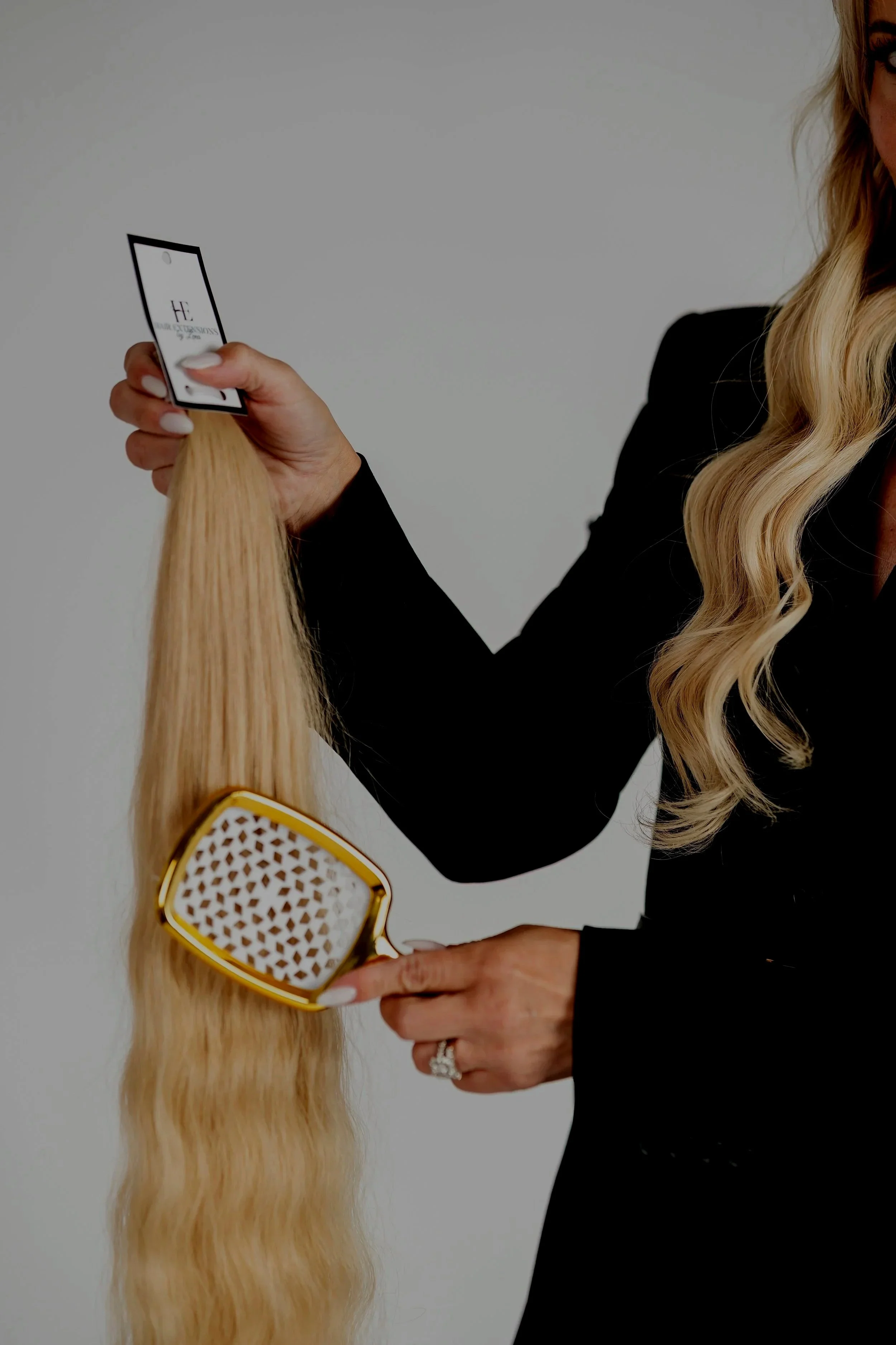 A woman with long blonde hair, dressed in black, is holding a card in one hand and a hairbrush in the other. She is brushing her long hair.