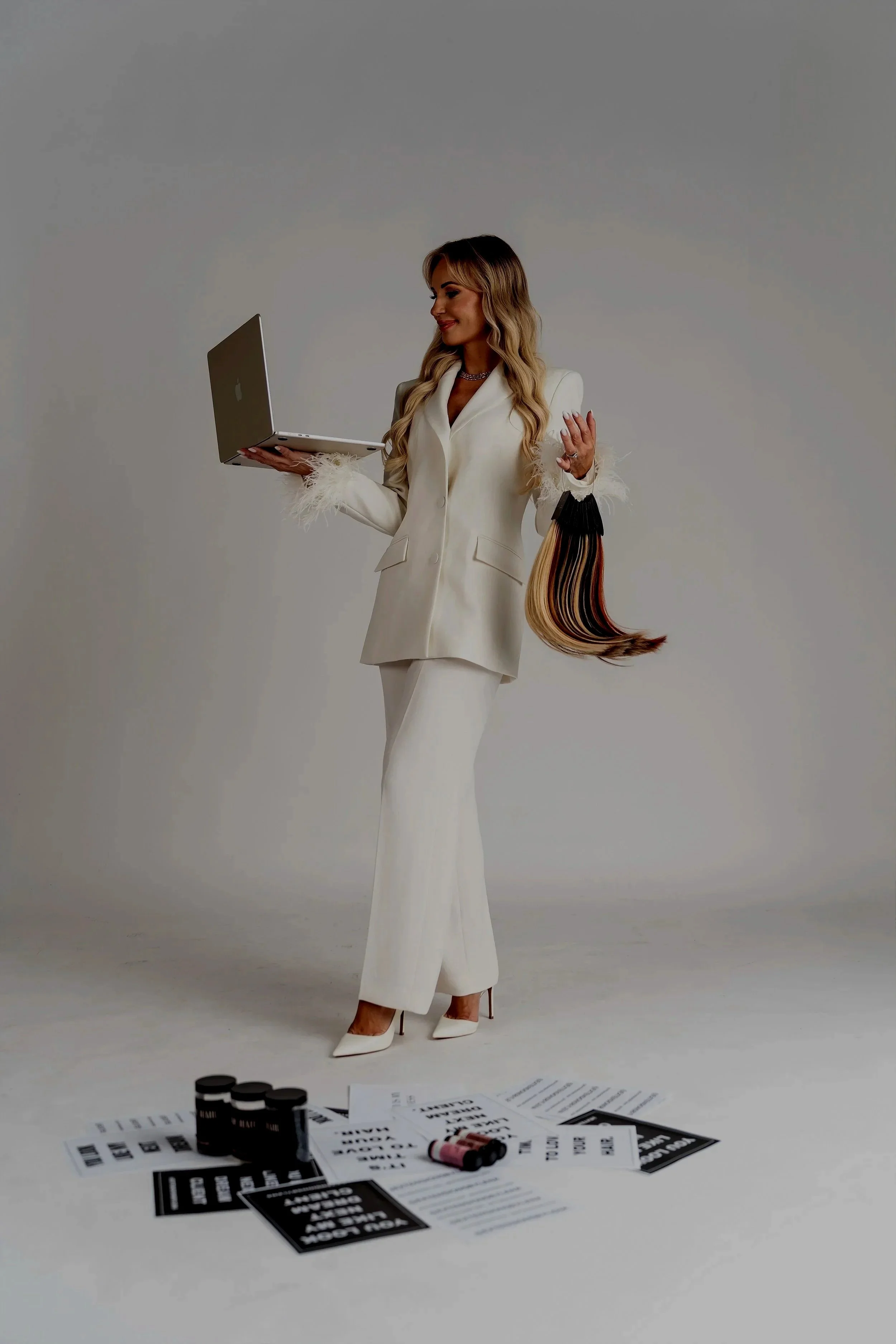 A woman in a white blazer and pants holding a laptop with a pile of papers and black coffee cups on the floor in front of her, against a plain gray background.