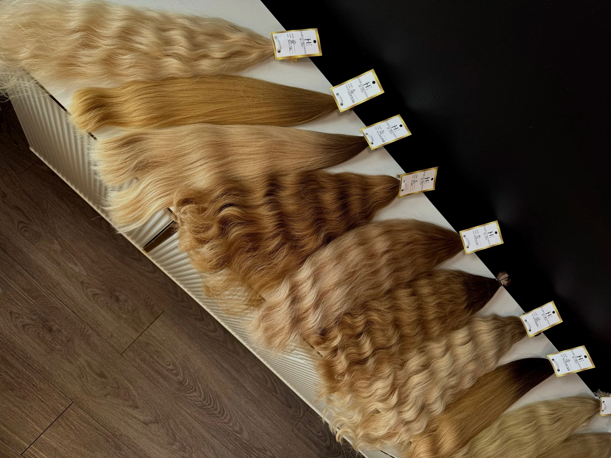 Display of four blonde hair extensions with wavy and straight textures, each labeled with tags showing product information and branding, arranged on a white and black surface.