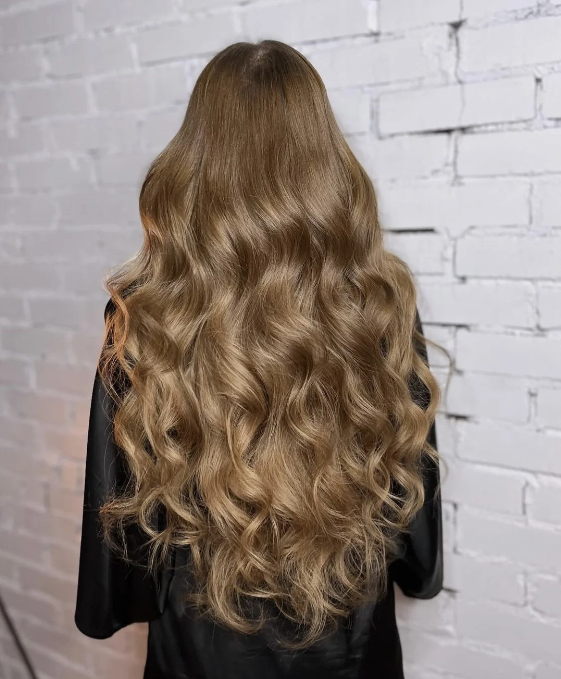 Back view of a woman with long, wavy, light brown hair against a white brick wall.