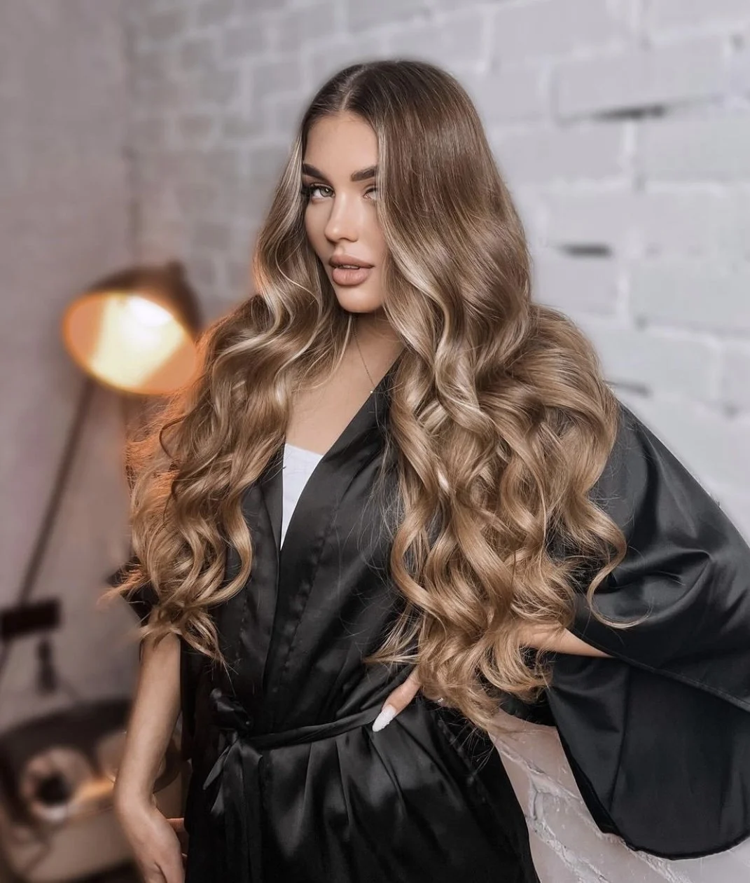 Young woman with long, wavy, light brown hair standing indoors against a white brick wall, wearing a black salon cape, with hairdresser tools and a lamp in the background.