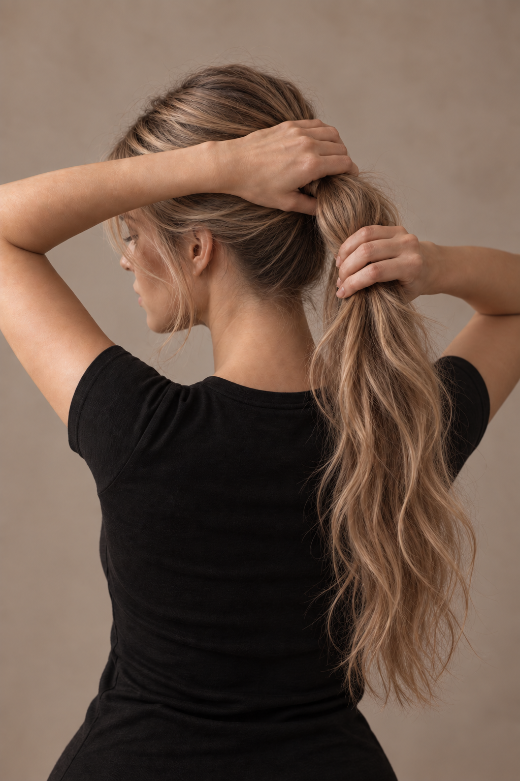 A woman with long, wavy, blonde hair in a ponytail, wearing a black t-shirt, is holding her hair up with both hands, facing sideways against a neutral background.