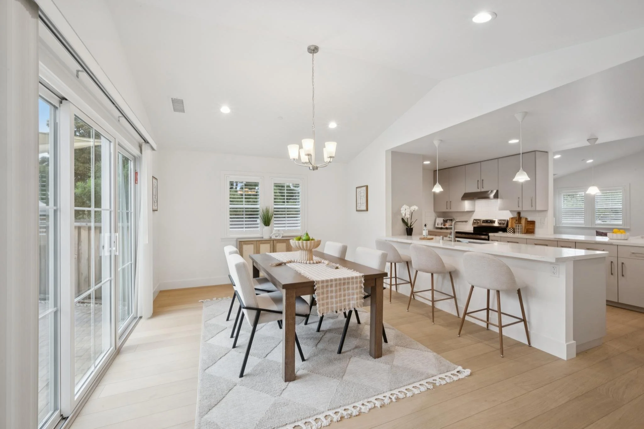 Bright dining and kitchen area with large sliding glass door, wooden dining table with six chairs, chandelier, white cabinets, kitchen appliances, and pendant lights.