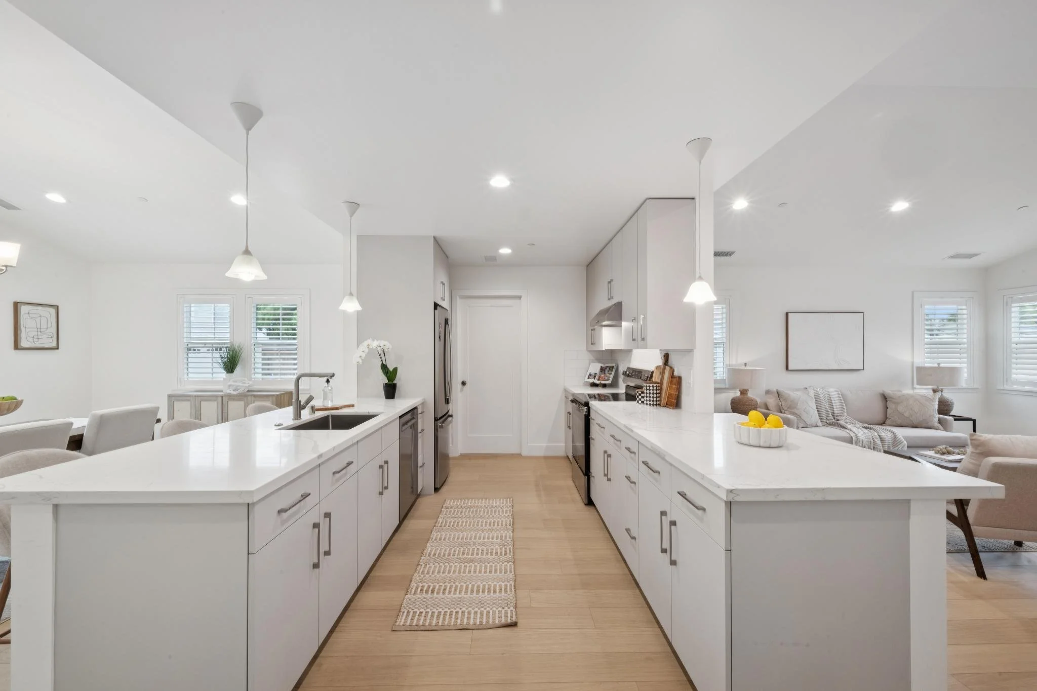 Bright white kitchen with island counters, pendant lights, and a view into a living room with sofa and windows.