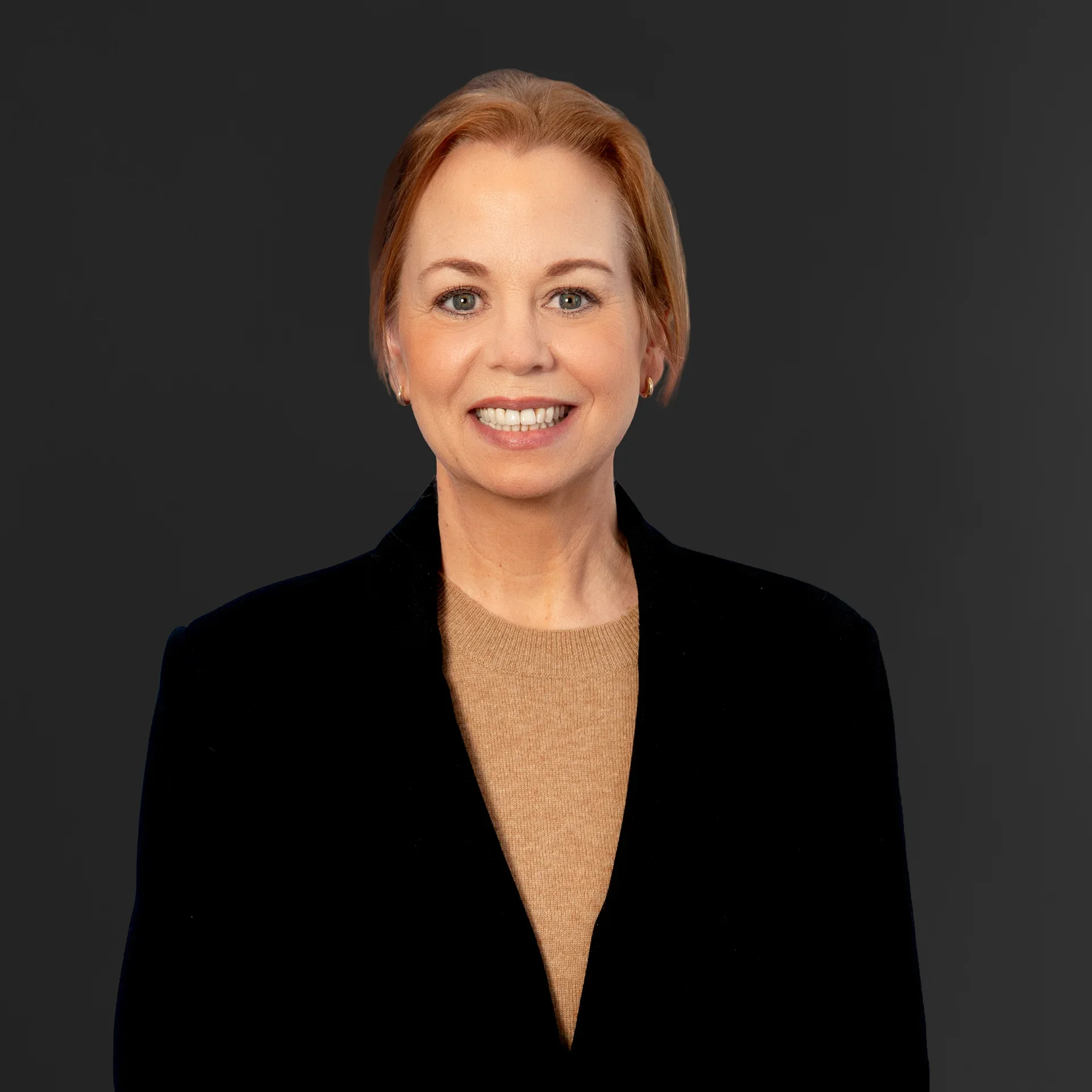A woman with short red hair smiling, wearing a black blazer and a tan shirt, against a dark gray background.