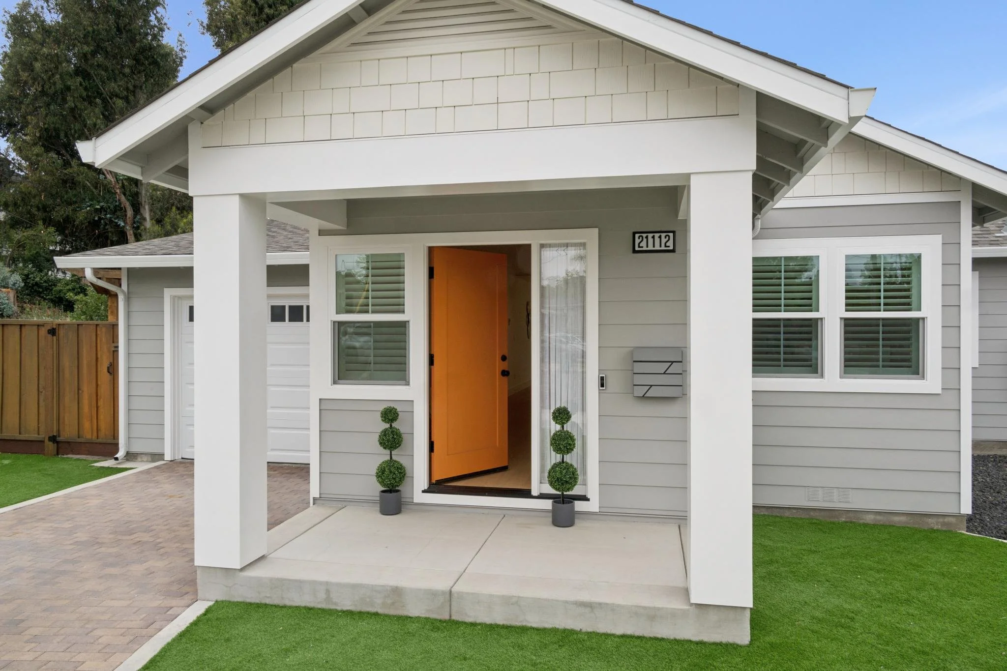 Front view of a modern house with a covered porch, gray siding, a bright orange front door, and decorative potted topiary plants, with a small lawn and a paved walkway.