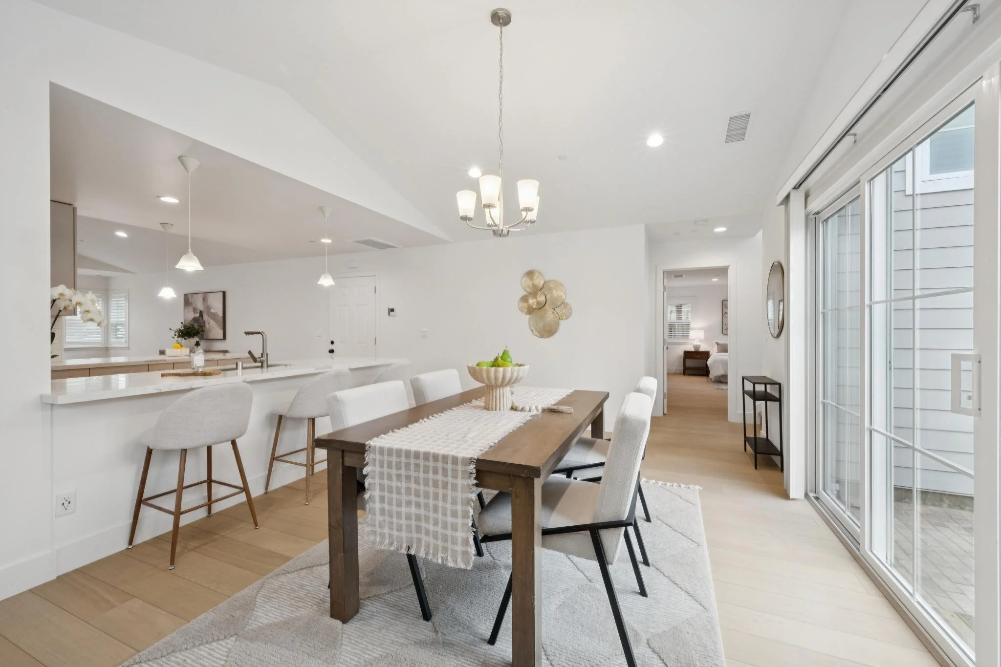 A spacious, modern dining area with a wooden table, six upholstered chairs, and a white table runner. There is a bowl of green pears on the table, and a large window with sliding glass doors on the right. The room features white walls, light wood flo