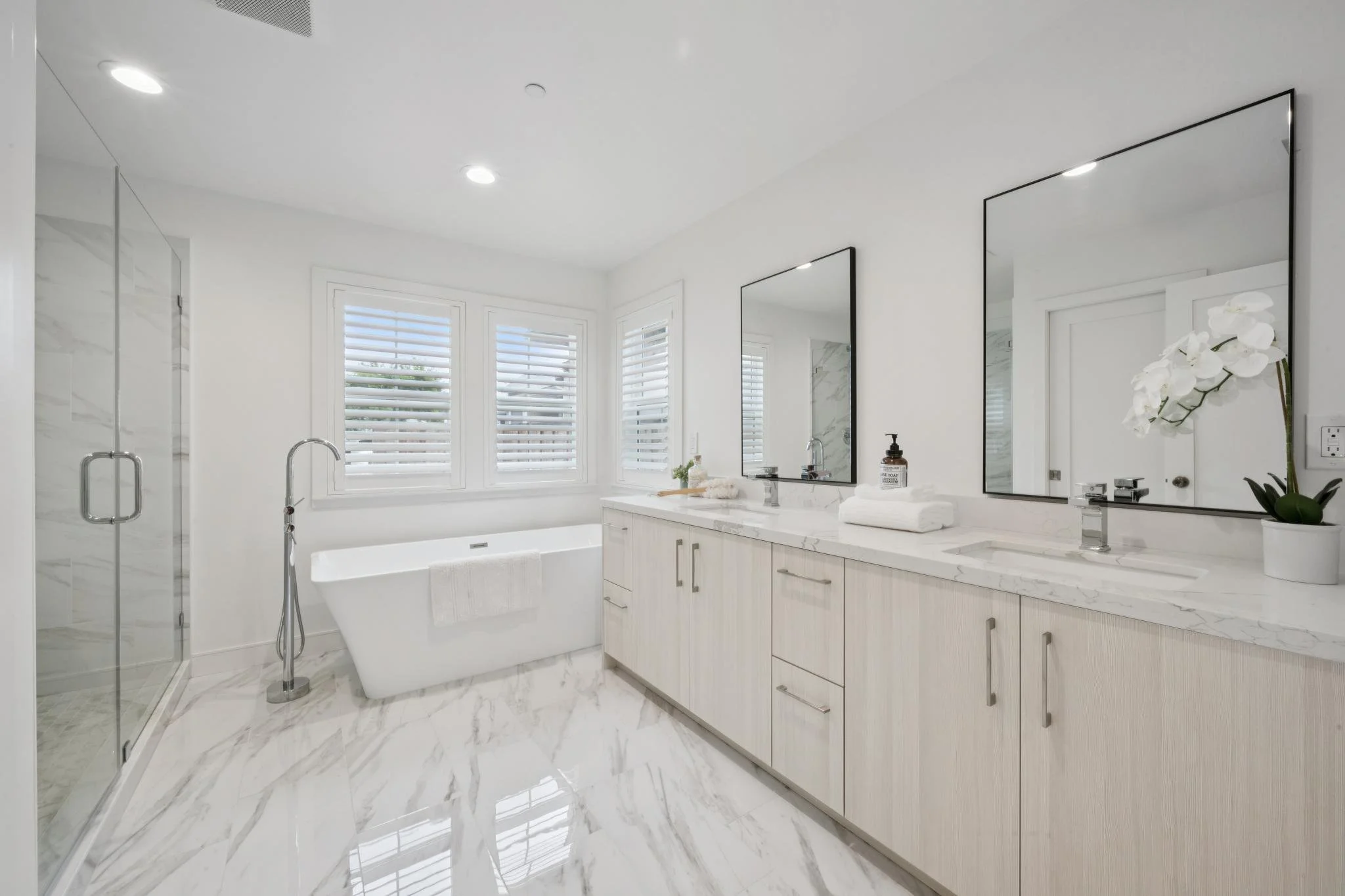 A modern, white bathroom with a large vanity, two mirrors, and a bathtub beneath three windows with white blinds. There is a glass shower enclosure on the left, and a small potted orchid on the right side of the vanity.