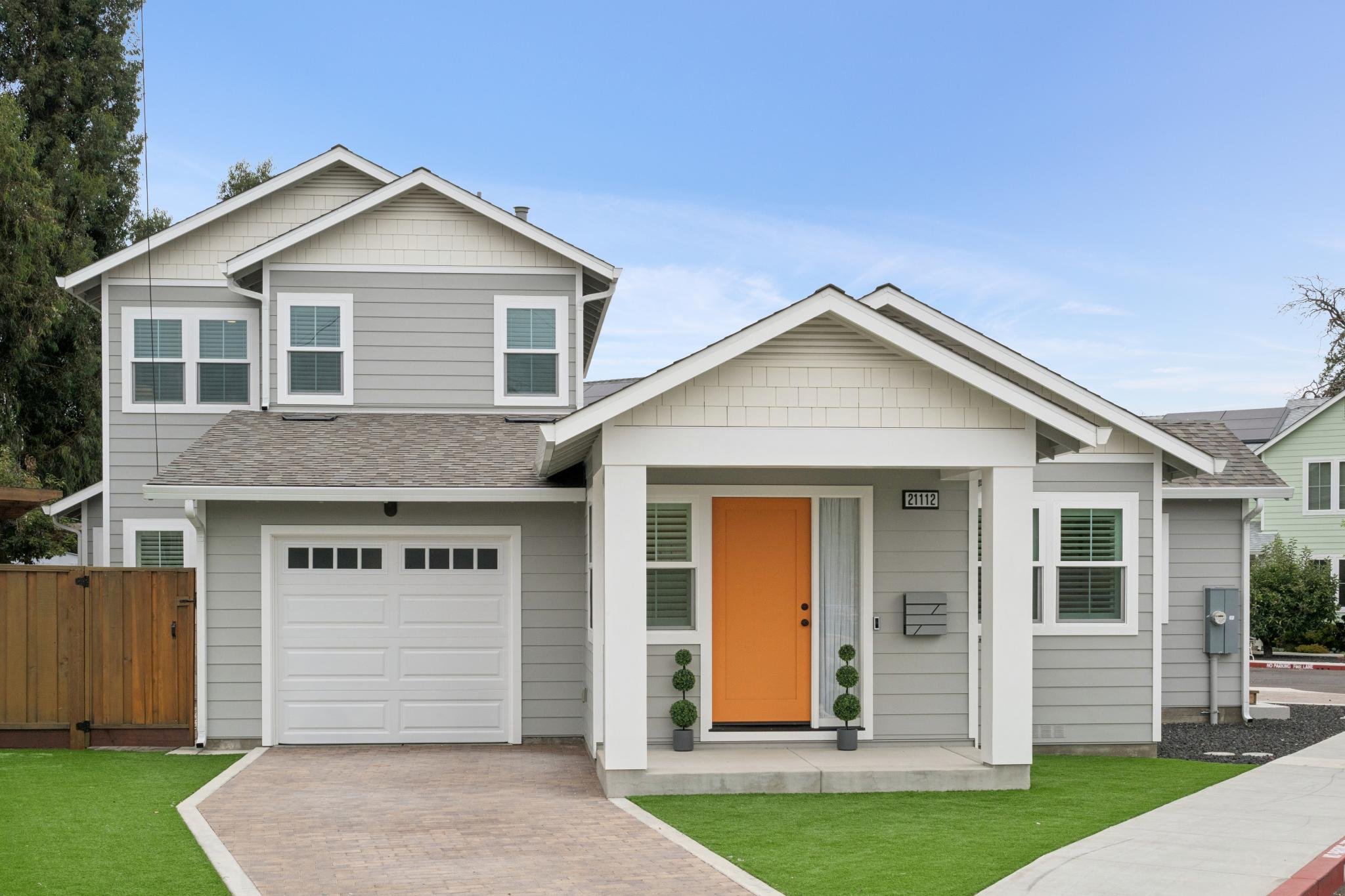 Front view of a modern two-story house with light gray horizontal siding and darker gray shingles, featuring a white garage door, an orange front door, and decorative bushes, with a well-maintained green lawn and a paved walkway leading to the entran