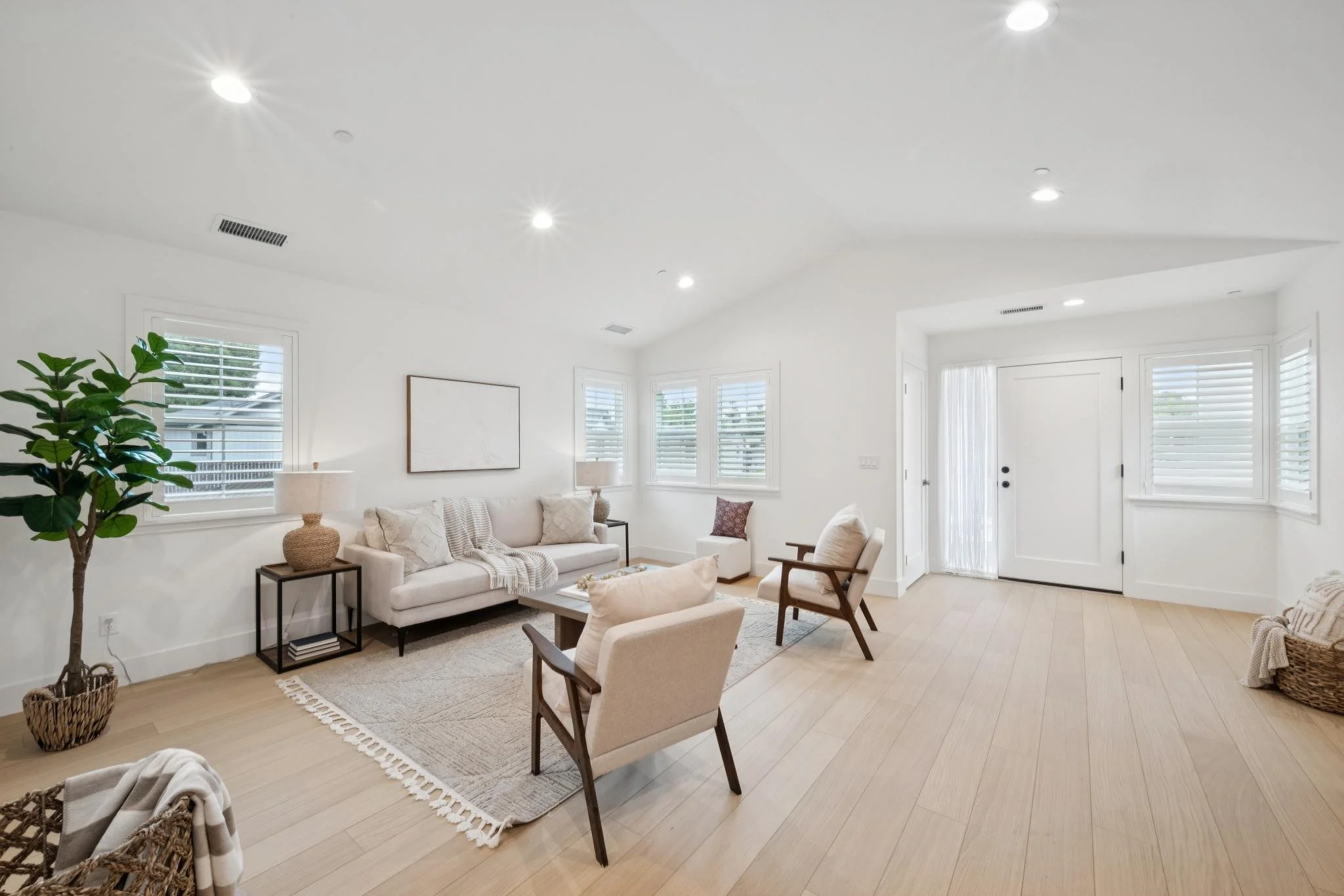 Bright living room with white walls, hardwood floors, a white couch with pillows, a wooden chair, a woven basket, and several windows with white blinds, illuminated by ceiling lights.