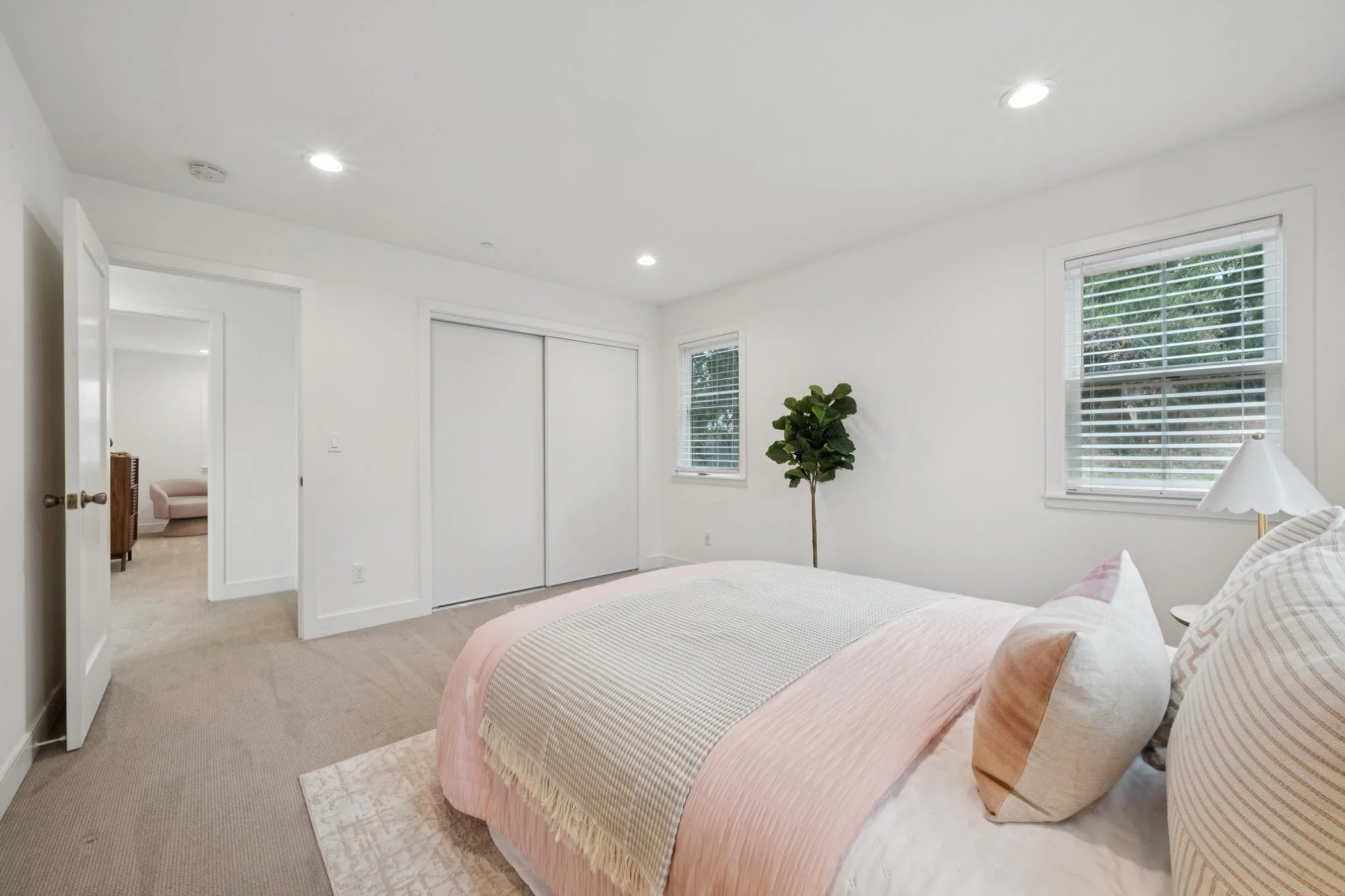 A bedroom with a bed covered in light pink and beige bedding, a white lamp, a green potted plant, and two windows with white blinds, with a doorway leading to another room.