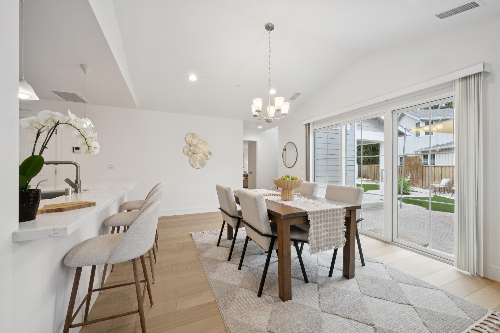 Bright, modern kitchen and dining area with large sliding glass door leading to backyard, white walls and ceiling, wooden dining table with six beige chairs, white orchid plant on kitchen counter, decorative wall art, circular mirror, chandelier ligh