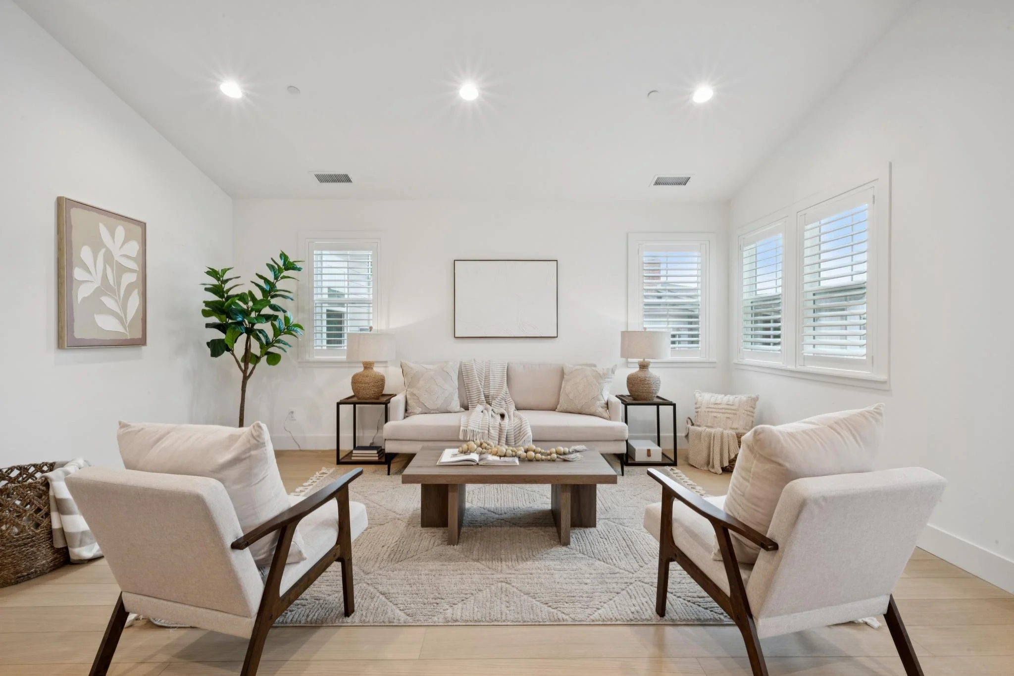 Bright living room with white walls, large windows with white shutters, beige sofa, armchairs, wooden coffee table with books and decor, potted plant, wall art, and floor lamps.