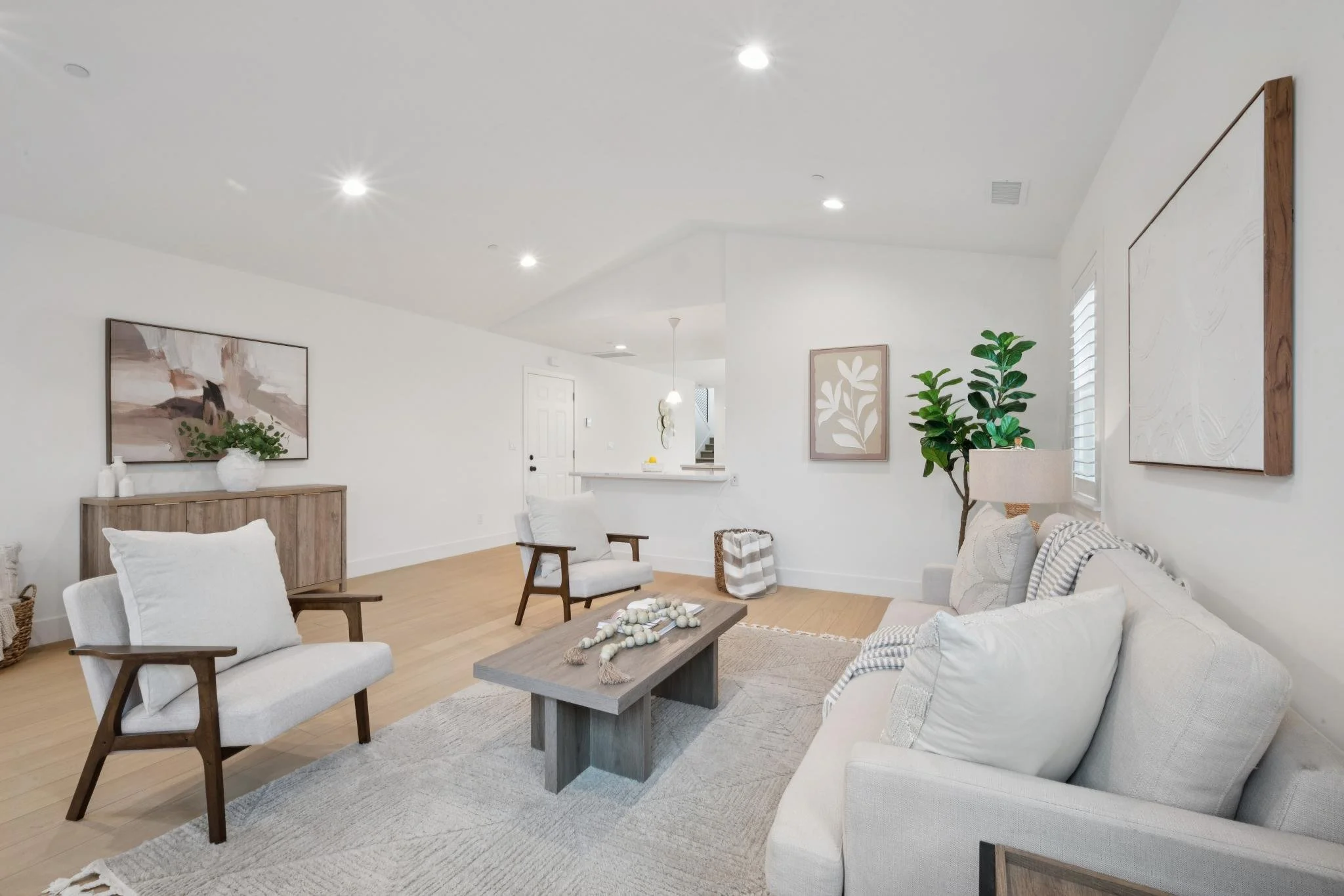 Bright living room with white walls, hardwood flooring, and modern furniture, including white sofas, wooden armchairs, and a rustic coffee table with decorative beads. Wall art, indoor plants, and a light fixture are visible.